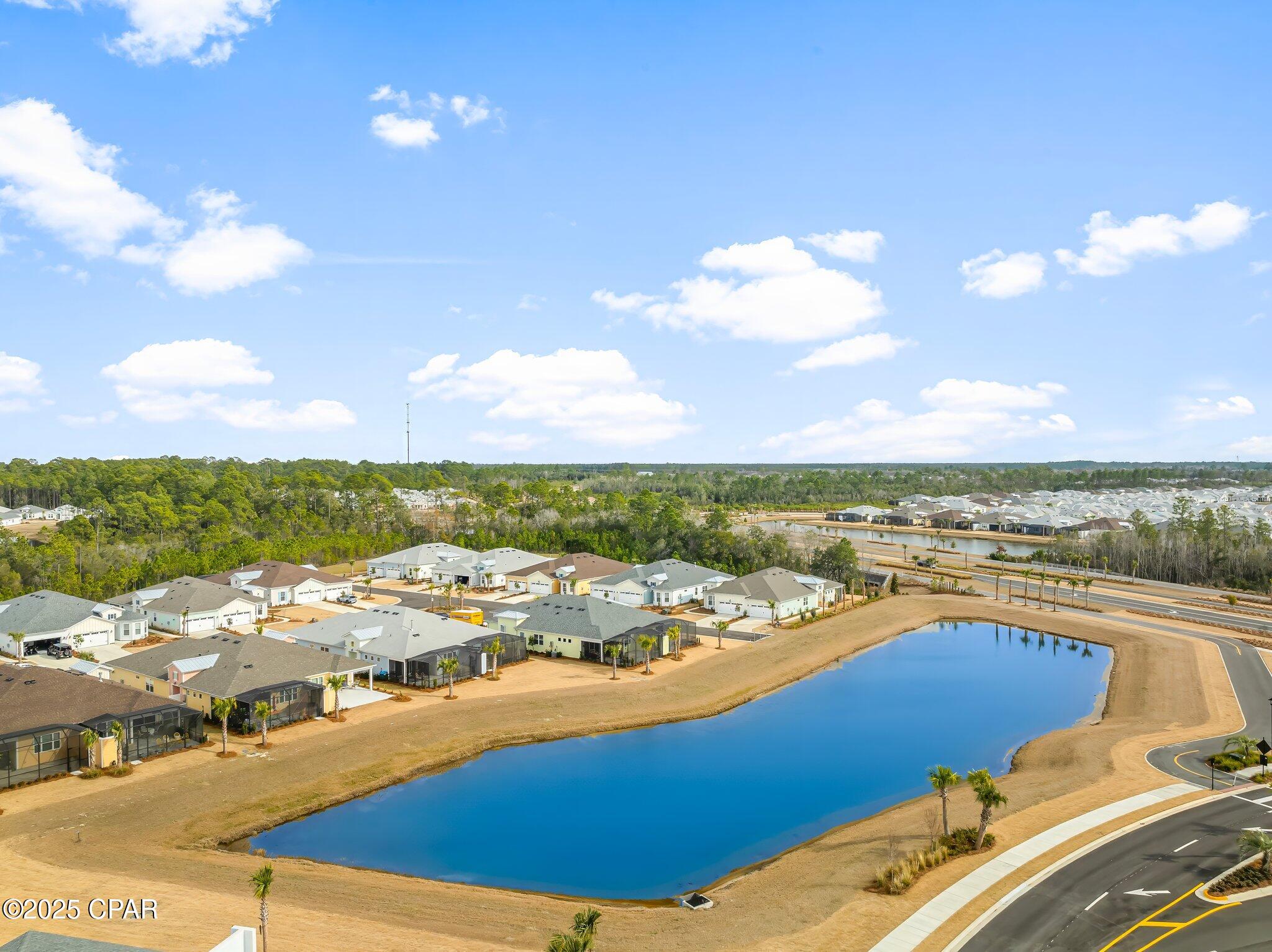 Aerial view of homes around a large pond, with wooded land and roads beyond.