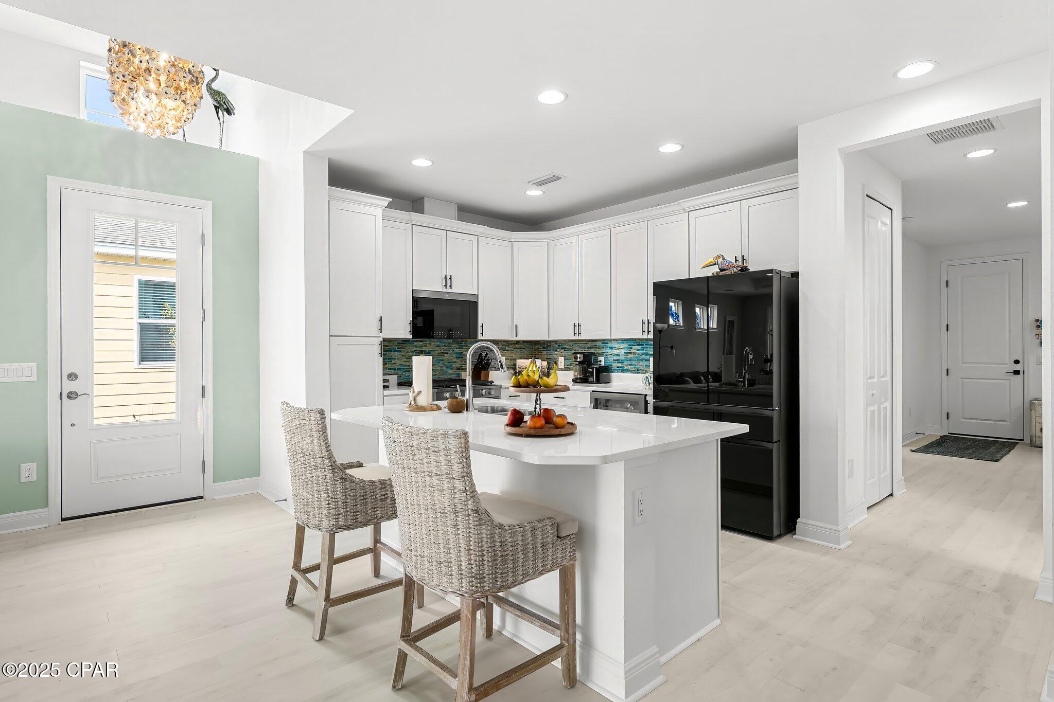Bright kitchen with white cabinets, colorful tile backsplash, black refrigerator, and a large island with woven barstools.