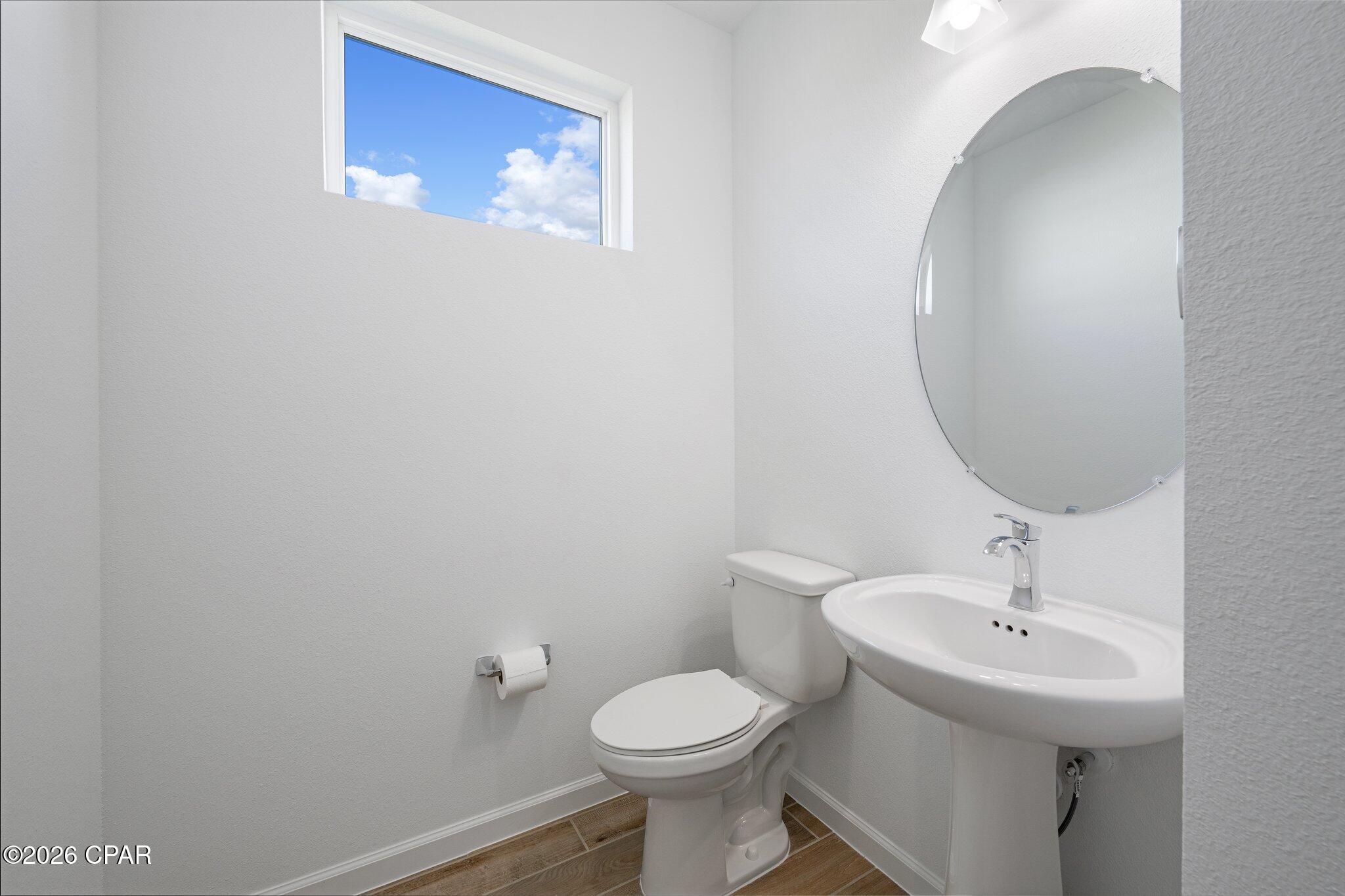 Half bathroom with a pedestal sink, round mirror, high window, and wood-look tile floor.
