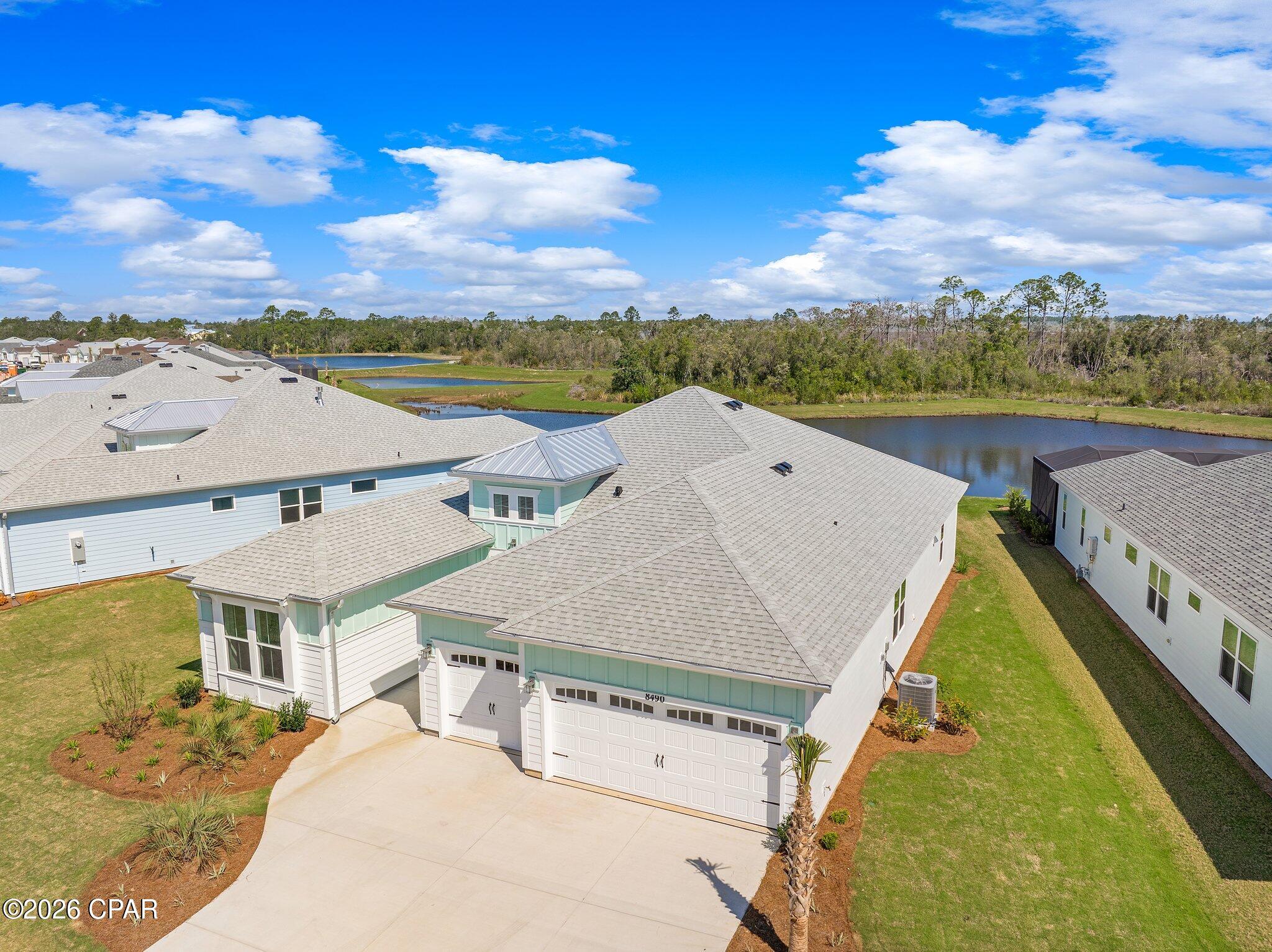 Aerial view of a white and aqua home beside a pond, with a broad driveway and gray roof.