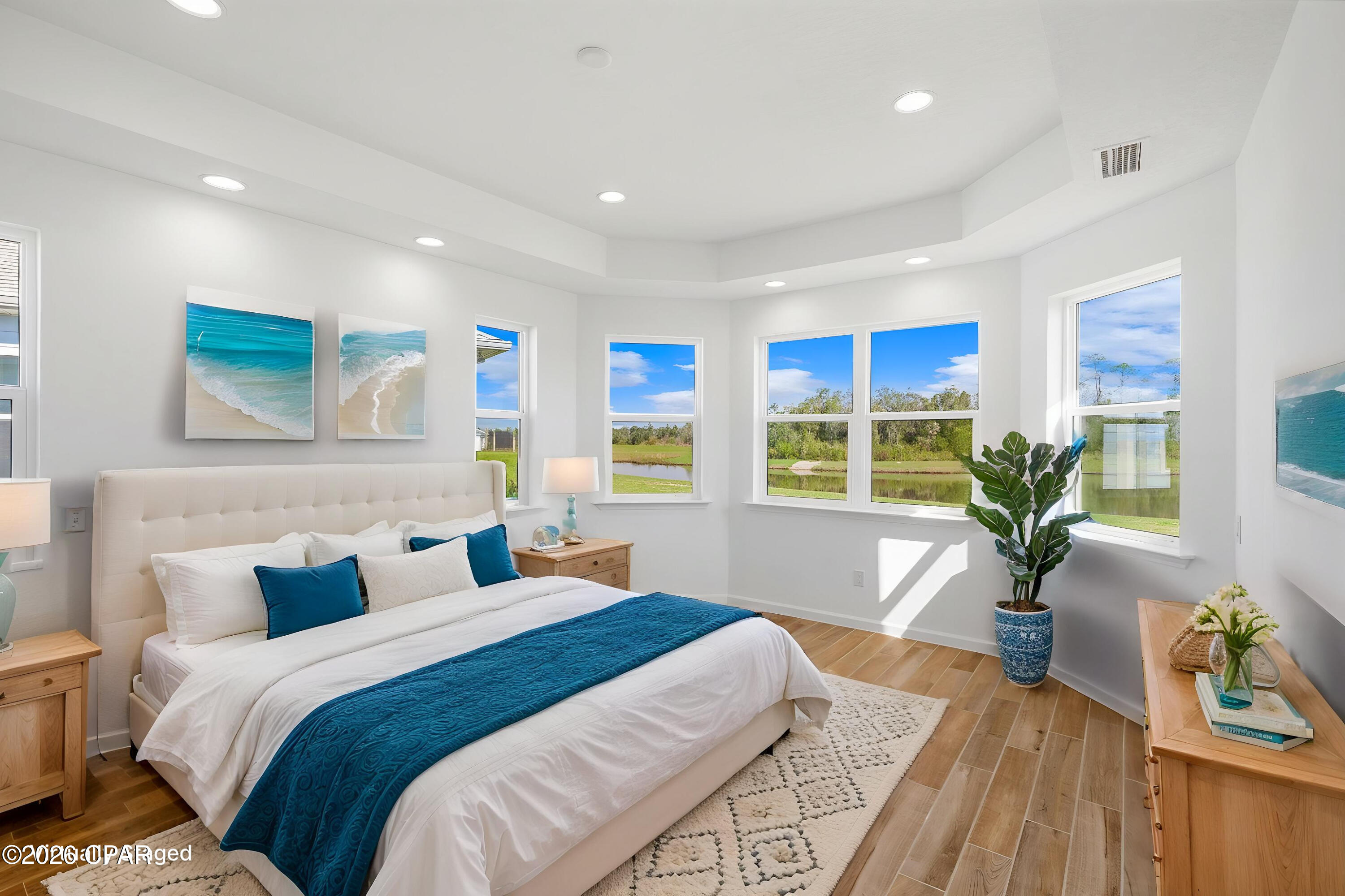 Bright bedroom with teal bedding, wood floors, and windows overlooking a pond.