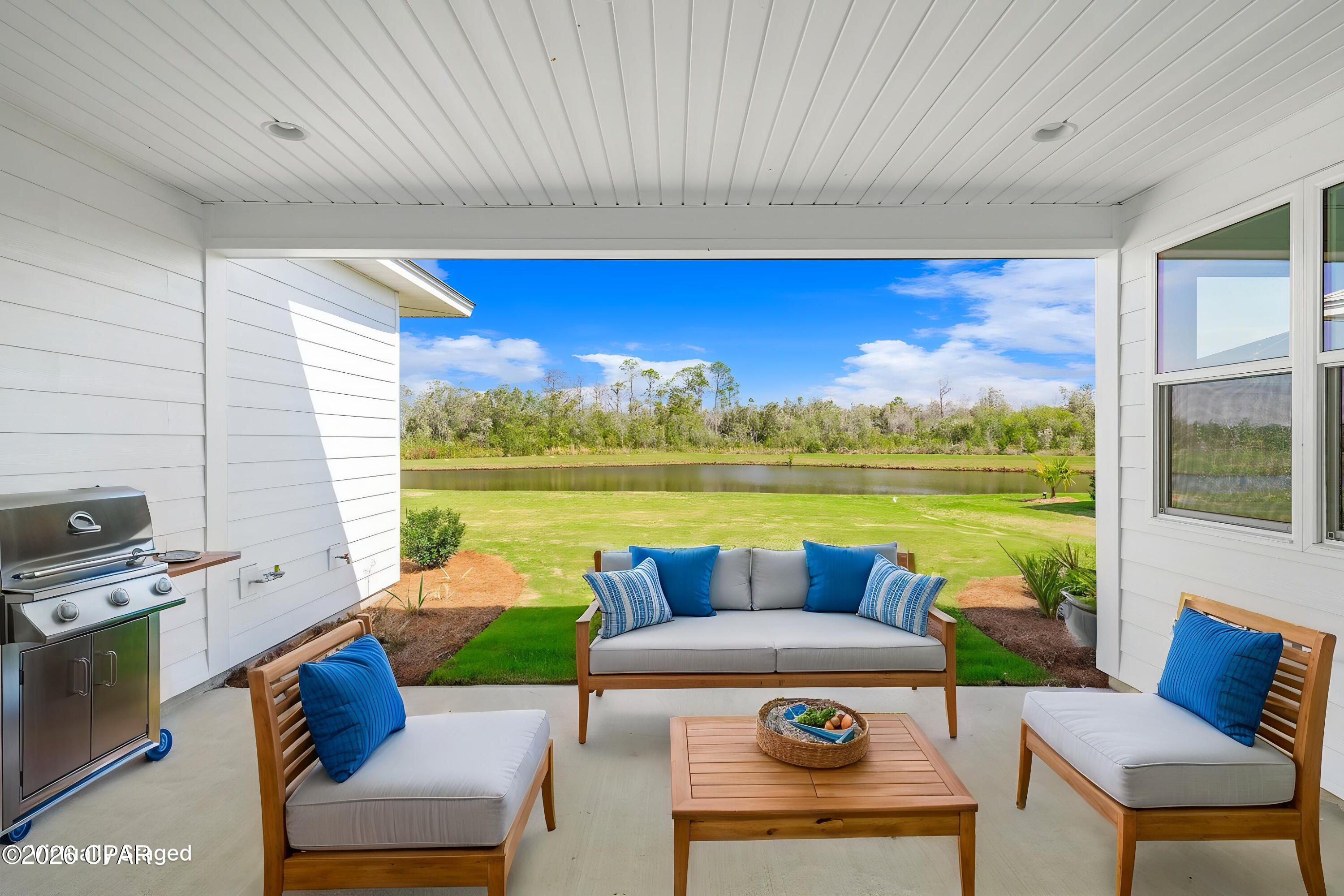 Covered patio with cushioned seating and a grill overlooking a pond and lawn.