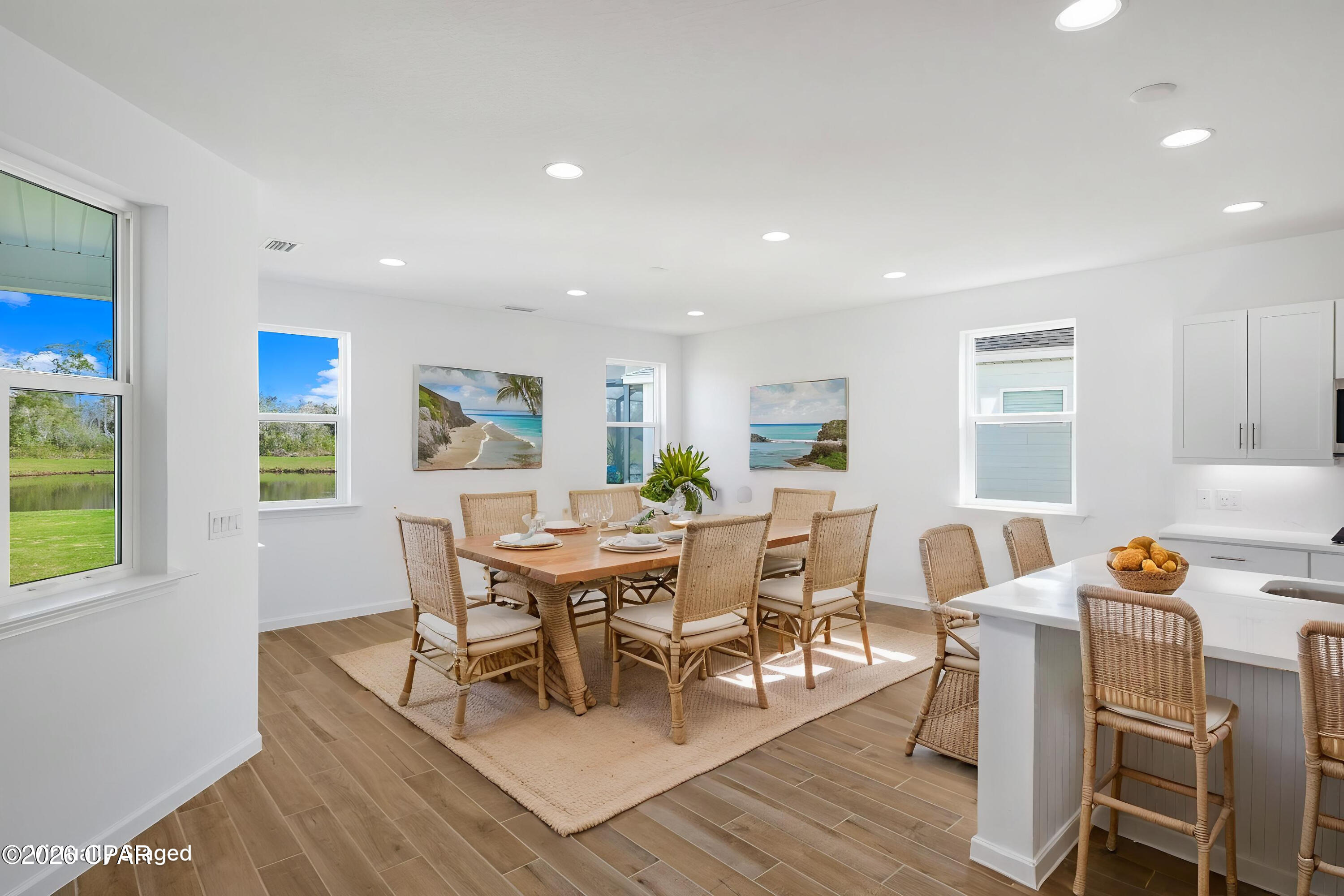 Dining area with a wooden table, woven chairs, coastal artwork, and a view of greenery through large windows.