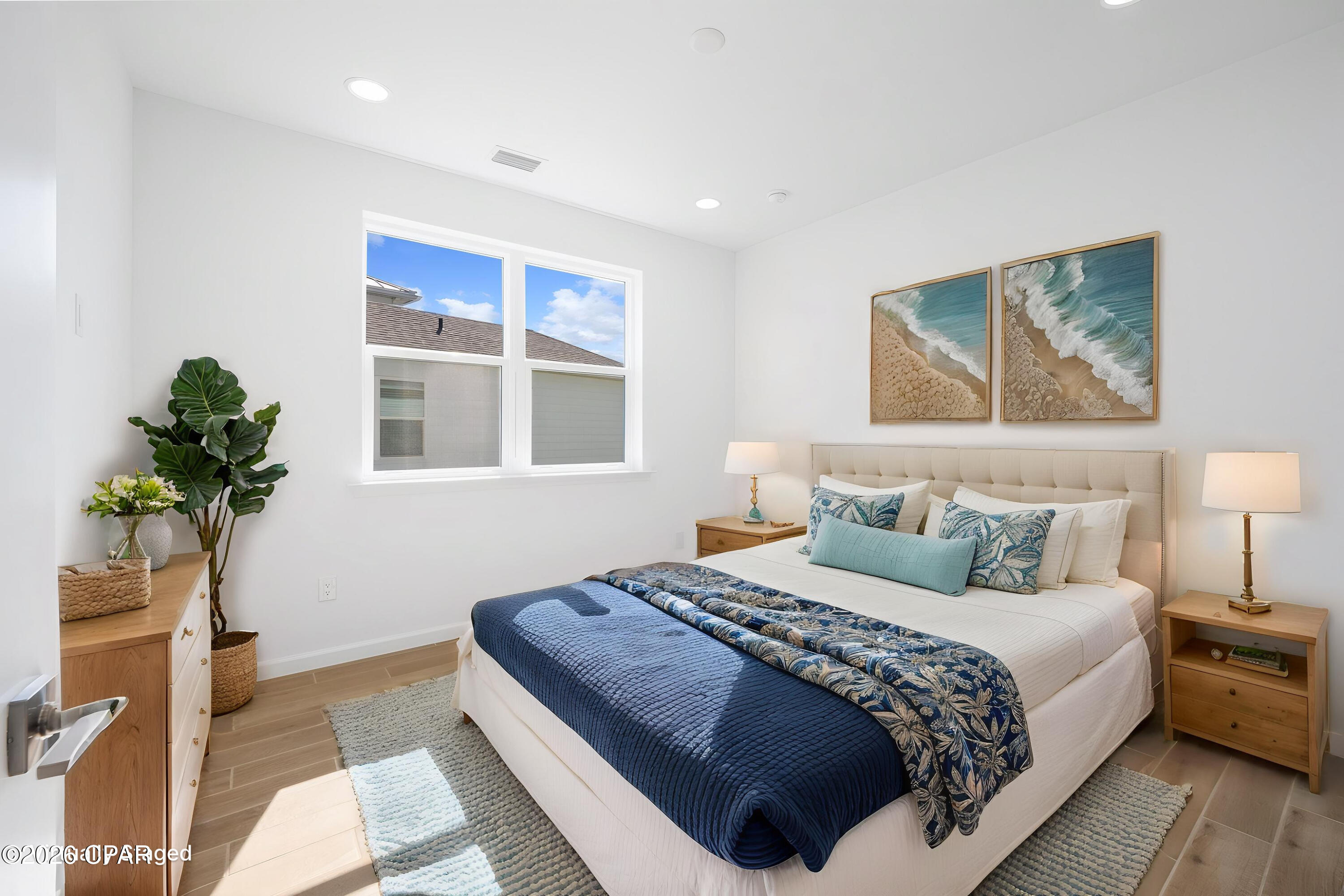Furnished bedroom with navy bedding, coastal artwork, and a window overlooking a neighboring roof.