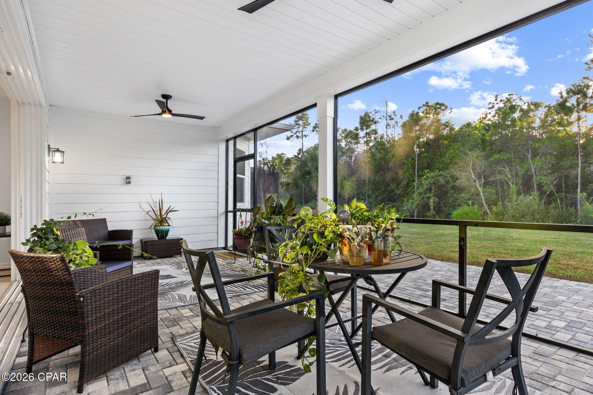 Covered patio with wicker seating, potted plants, and a wooded view beyond the lawn.