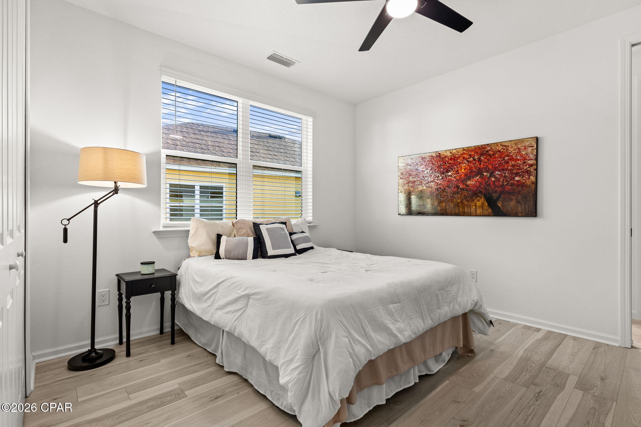Furnished bedroom with a white bed, large window, light wood floors, and autumn-tree artwork.