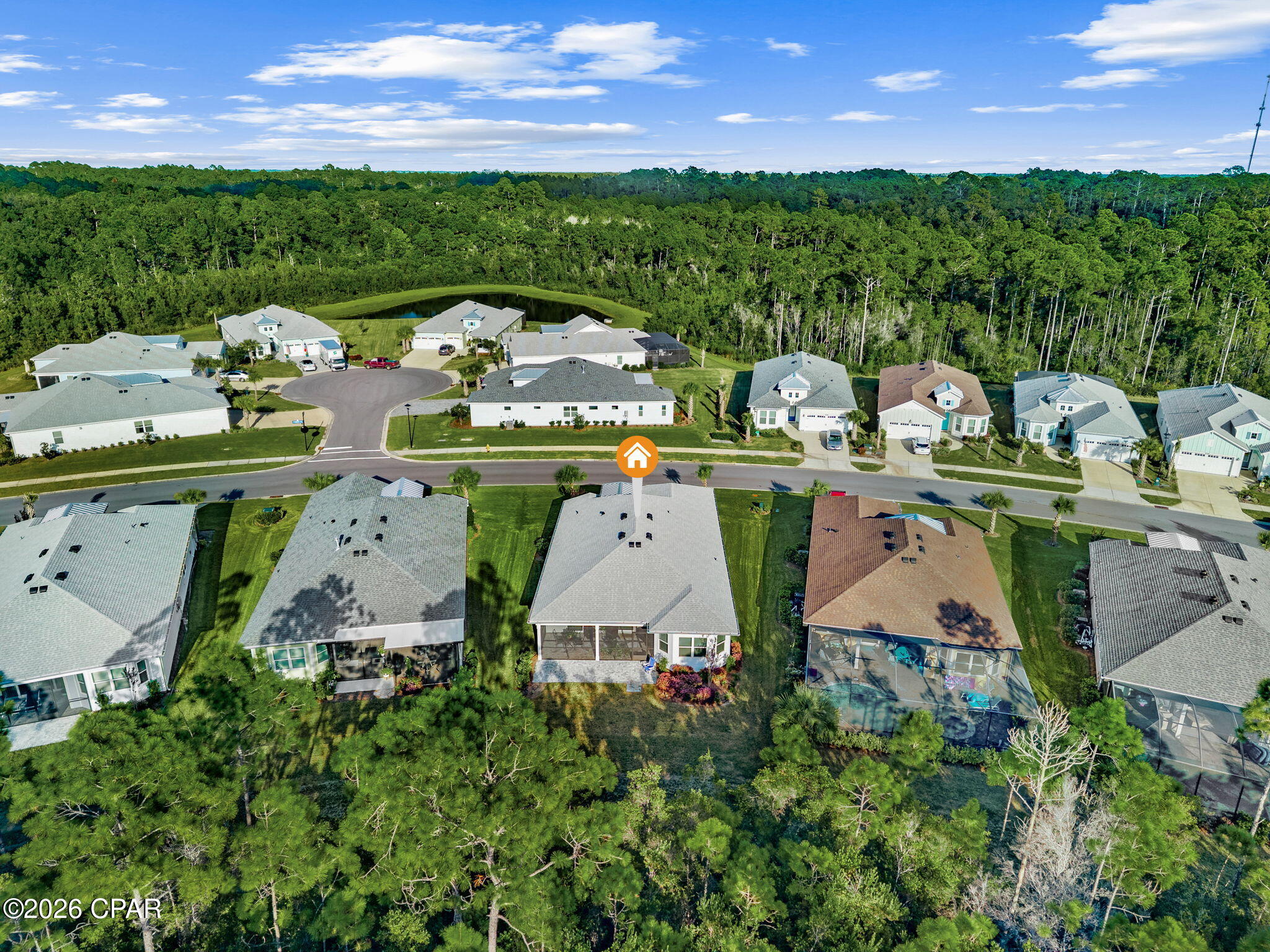 Aerial view of the marked single-story home, with a screened patio and dense woodland beyond the neighborhood.