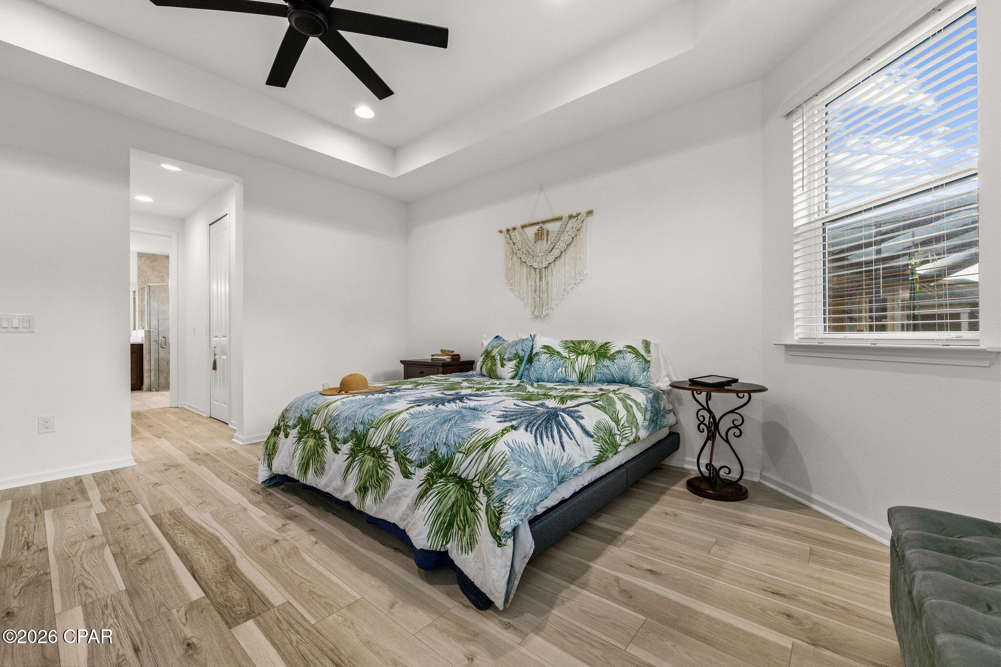 Bedroom with tropical-leaf bedding, light wood-look floors, and a window with white blinds.