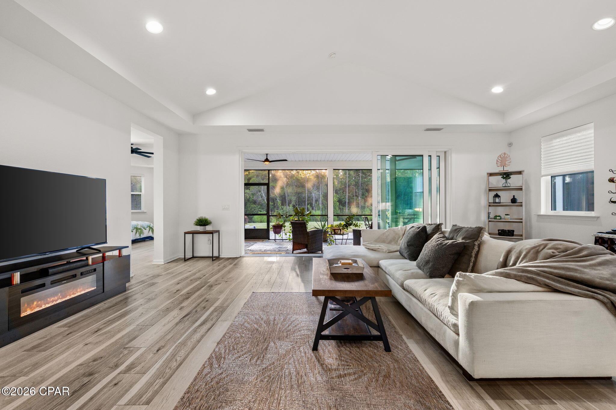 Living room with a vaulted ceiling, sectional sofa, electric fireplace, and sliding doors to a tree-lined patio.