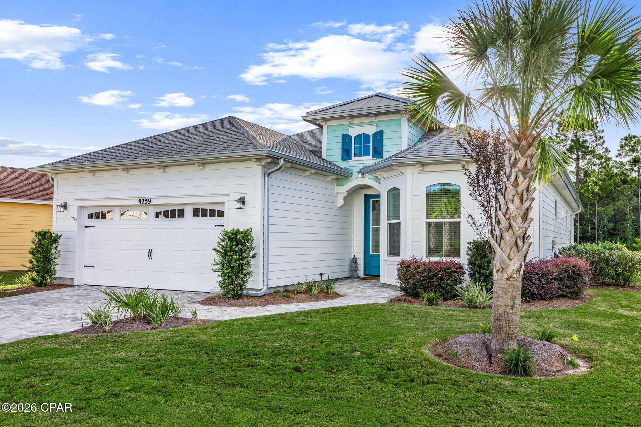 White coastal-style house with turquoise door, blue shutters, two-car garage, and a palm tree in the front yard.