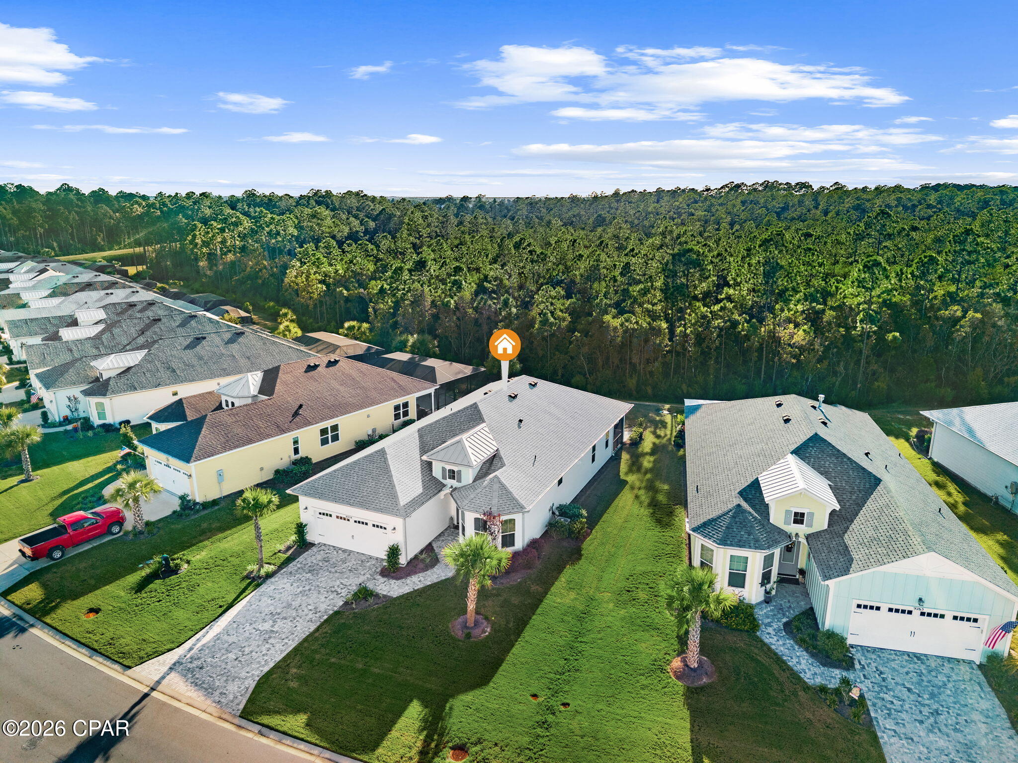 Aerial view of a marked single-story home beside a wooded area and neighboring houses.