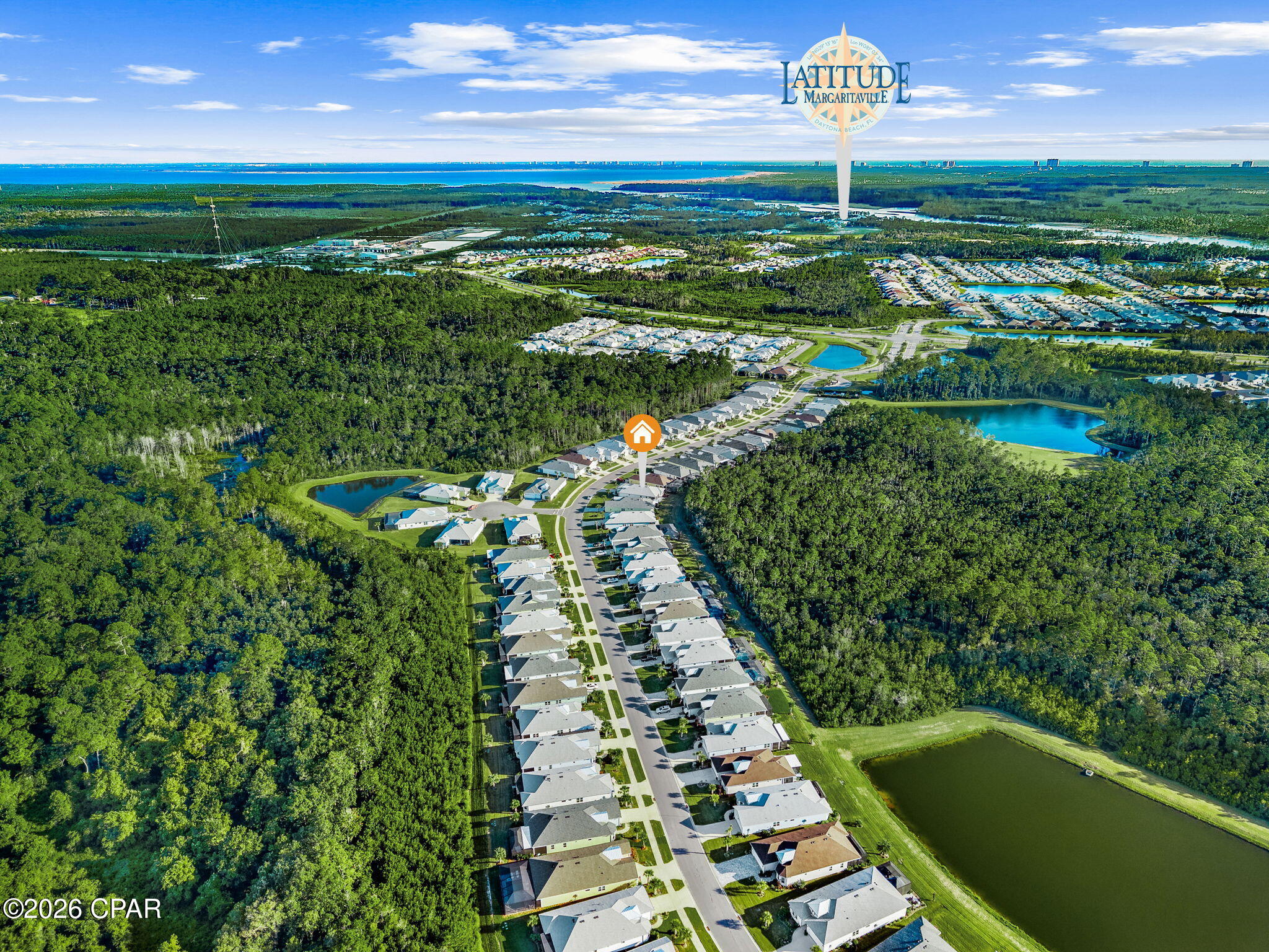 Aerial view of a coastal neighborhood, with a marked home, wooded areas, ponds, and ocean in the distance.