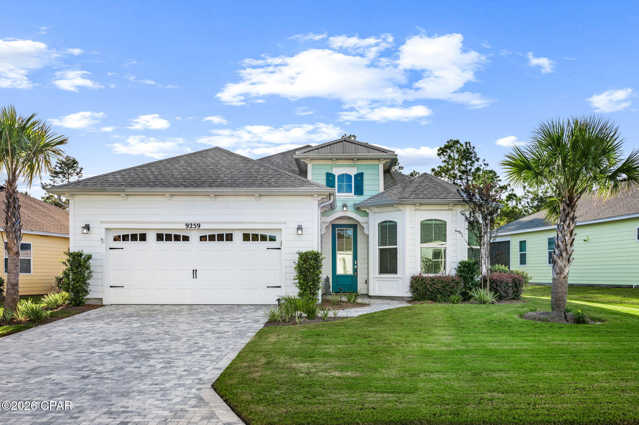 White coastal-style house with turquoise entry and shutters, two-car garage, paved drive, lawn, and palm trees.
