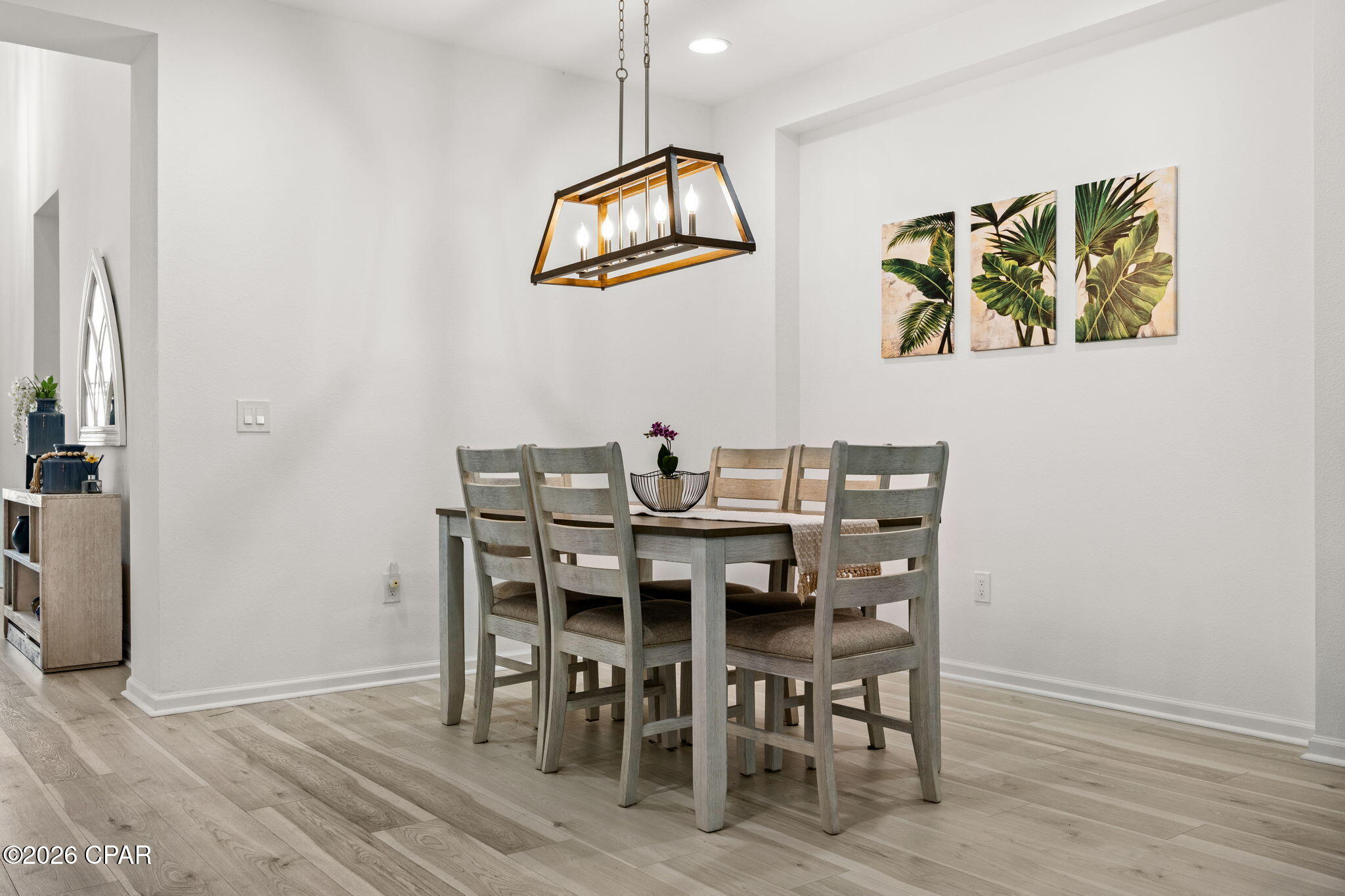 Dining room with a gray wood table, ladder-back chairs, geometric pendant light, and tropical leaf wall art.