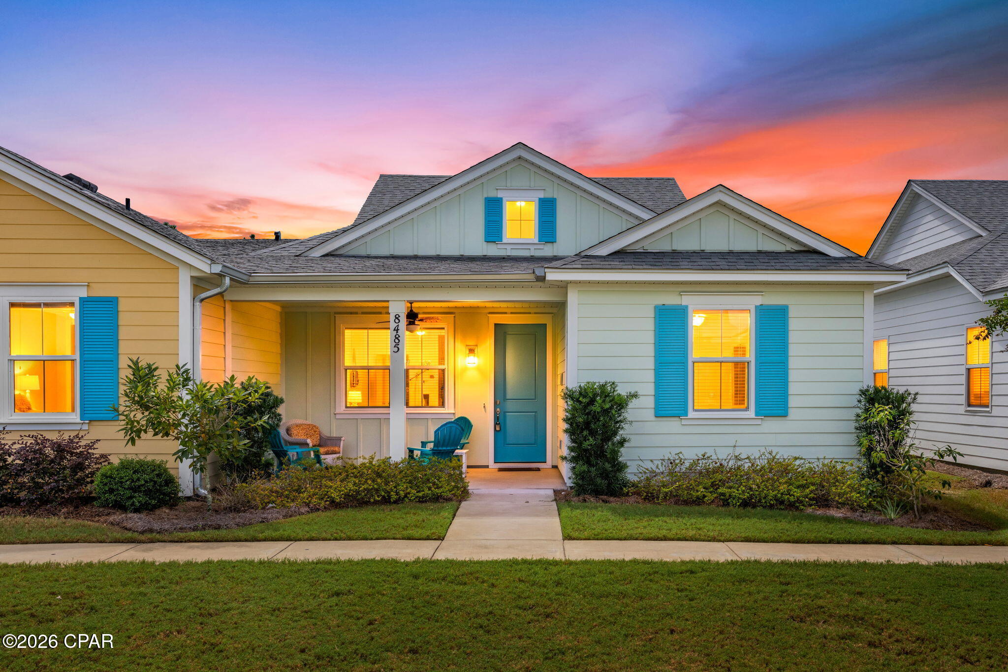 Single-story home with blue shutters and a covered entry, glowing windows, and landscaped front yard at dusk.