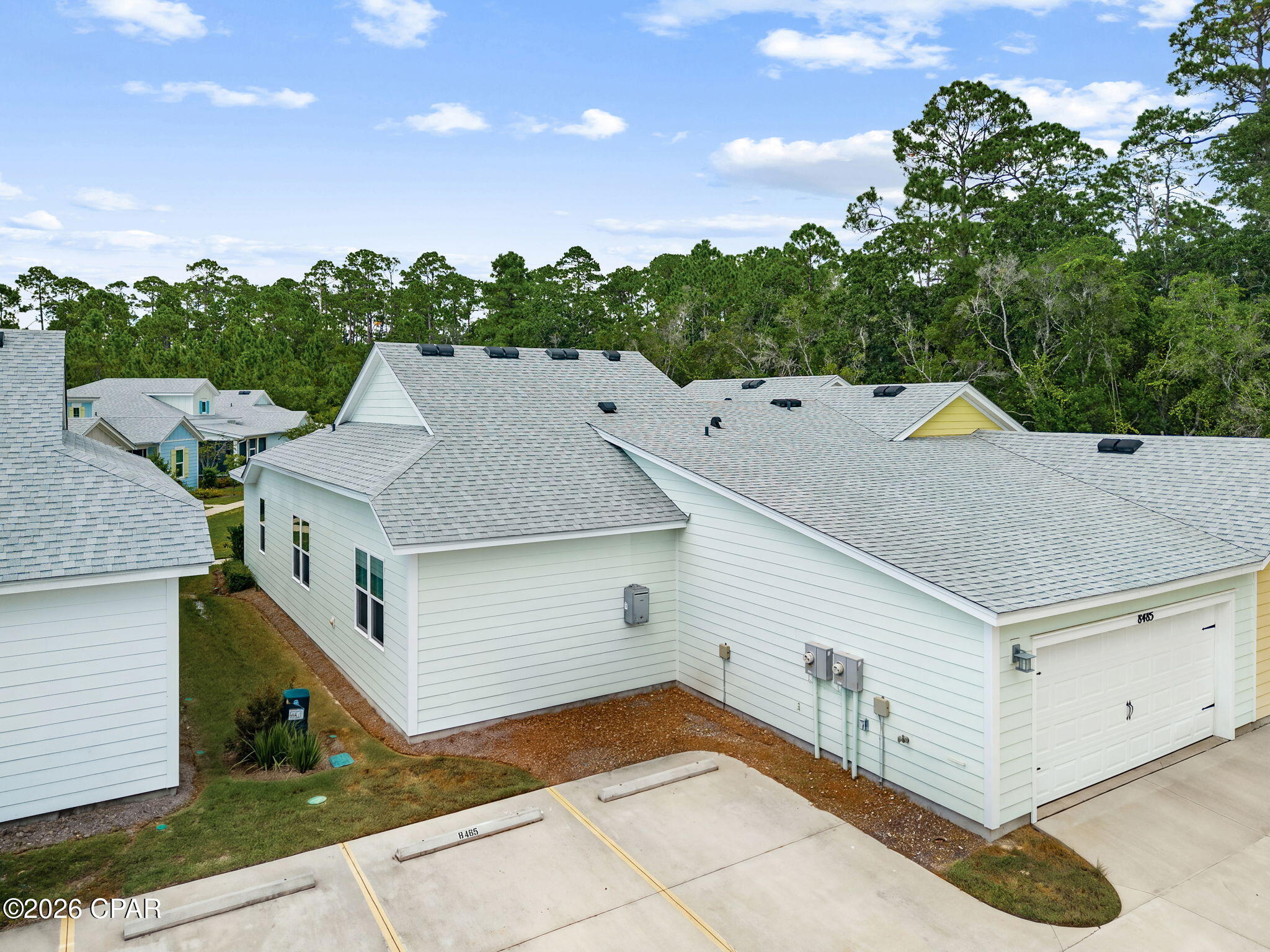 Elevated view of a white home with an attached garage, paved parking area, and woods behind it.
