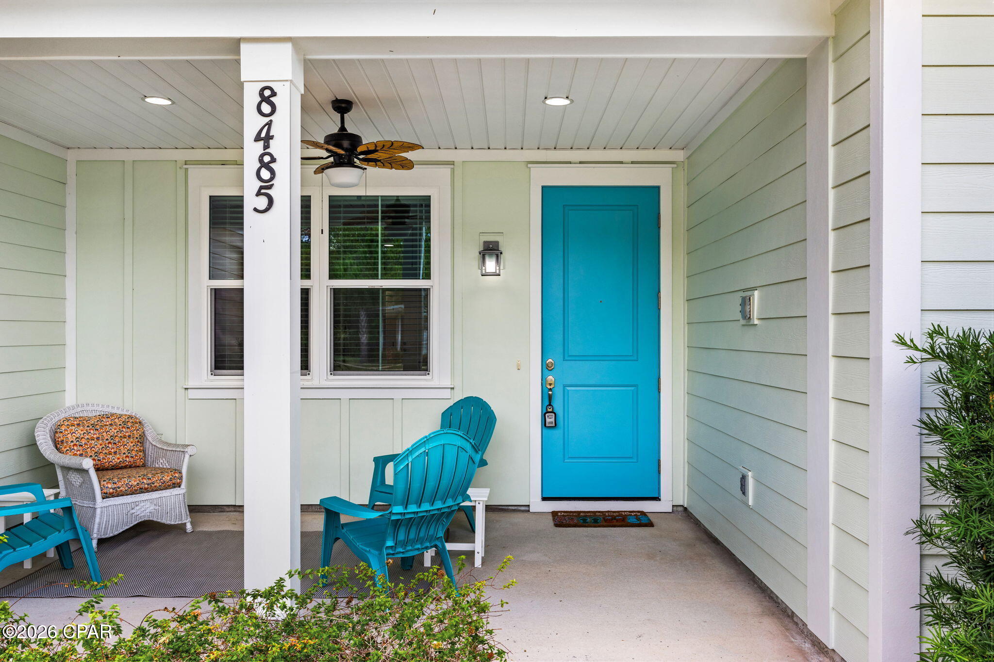 Covered front porch with a bright turquoise door, white-trimmed windows, and colorful outdoor chairs.