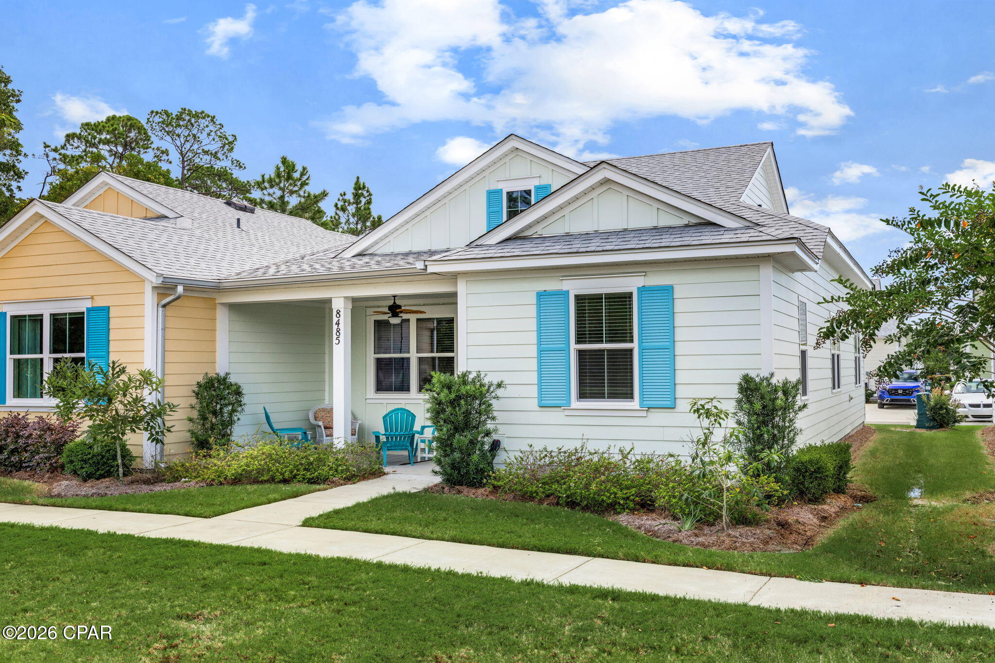 White-sided home with bright blue shutters, a covered porch, and turquoise chairs amid landscaped shrubs.