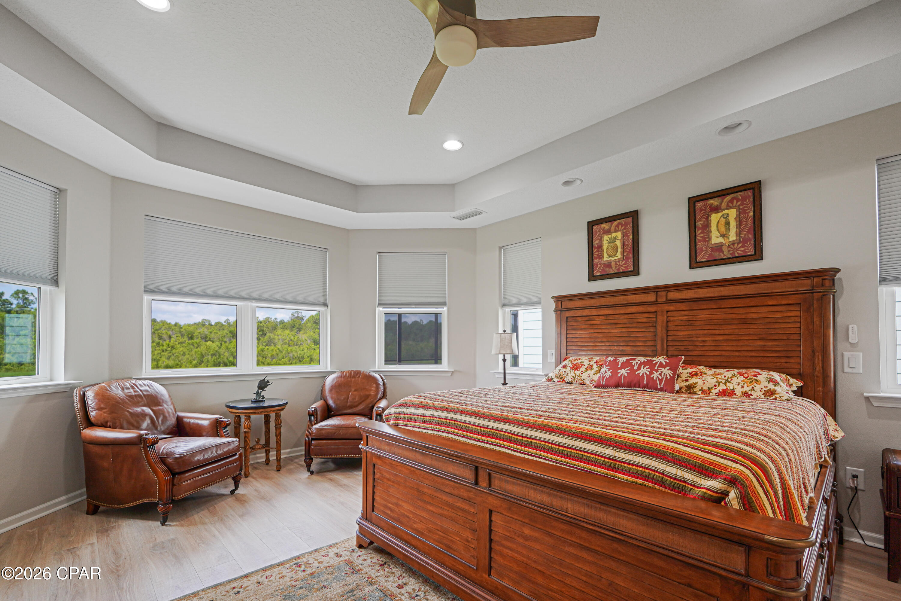 Bedroom with a large wooden bed, tropical bedding, leather armchairs, and windows overlooking trees.
