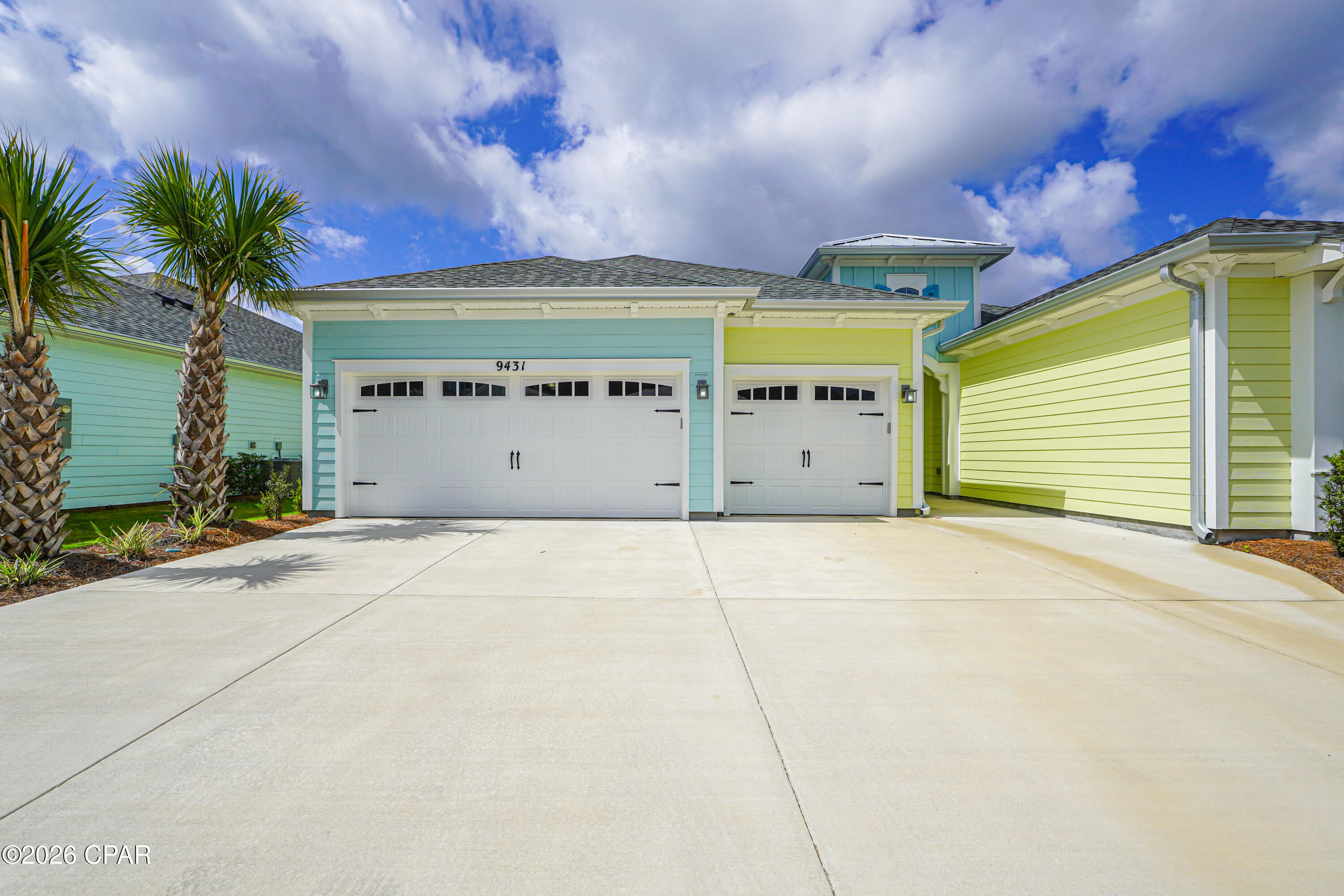 Blue and yellow coastal-style home with two garage doors and palm trees beside a wide driveway.