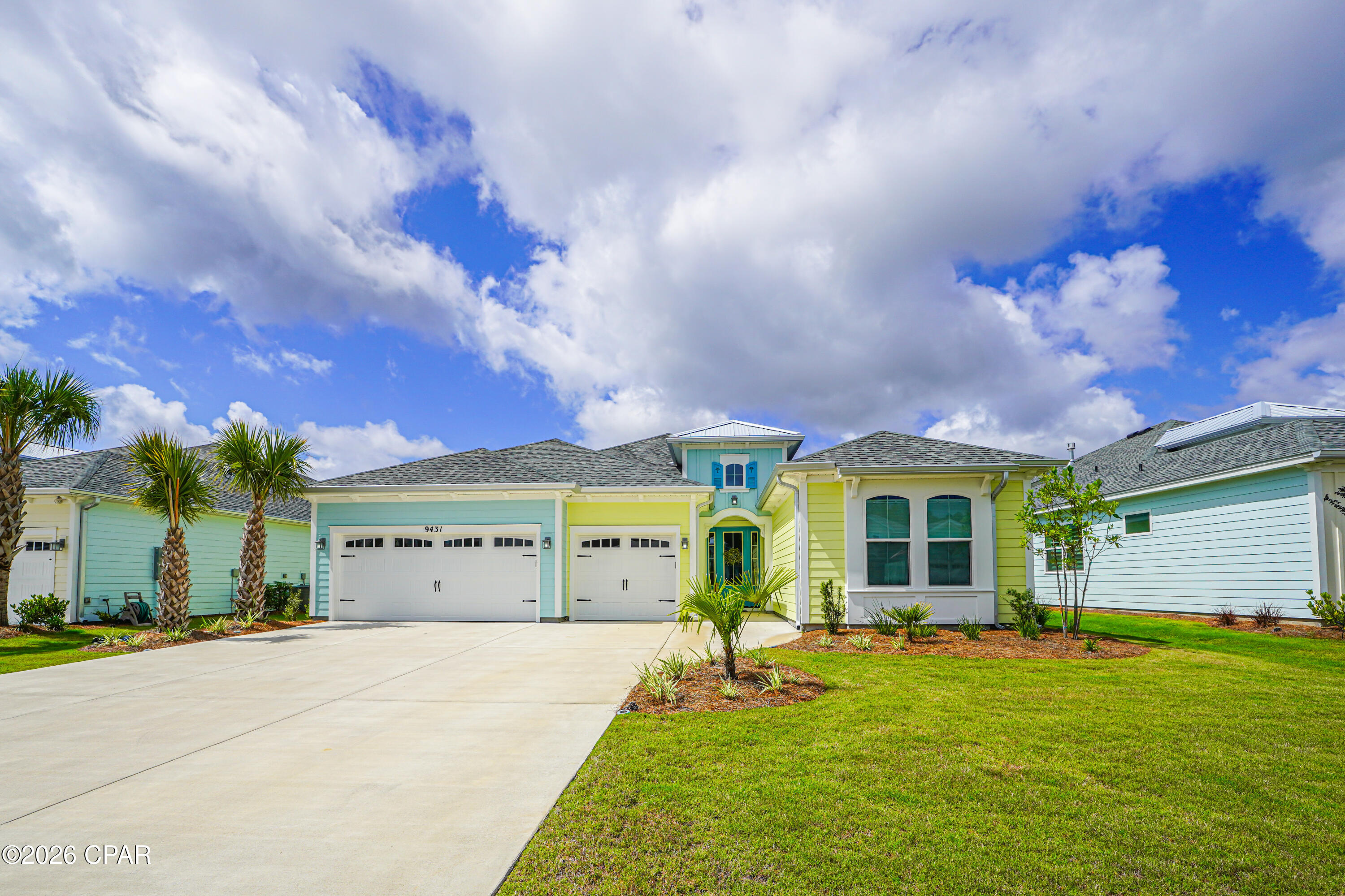 Coastal-style home with aqua and yellow siding, two garages, palm trees, and a broad driveway.