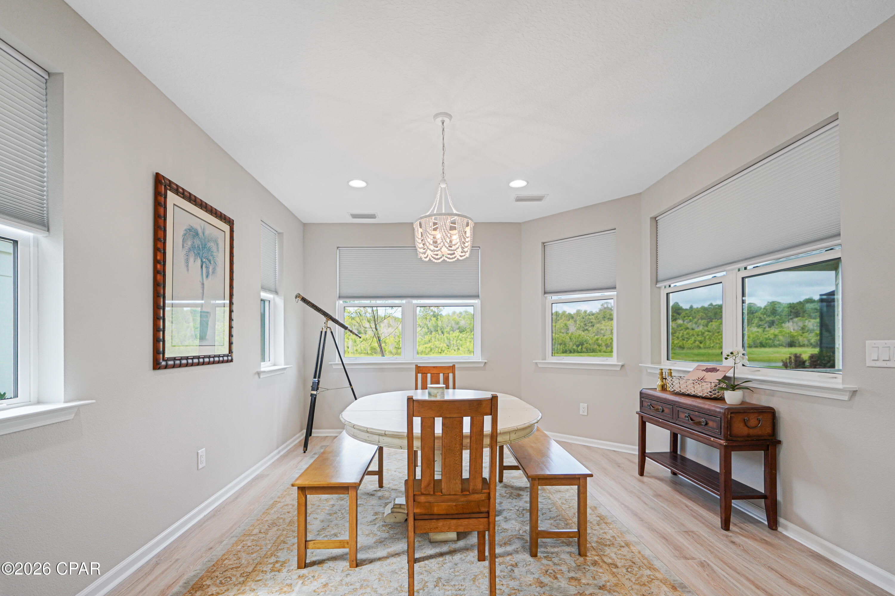 Dining room with a round table, wooden chairs, chandelier, and large windows overlooking greenery.