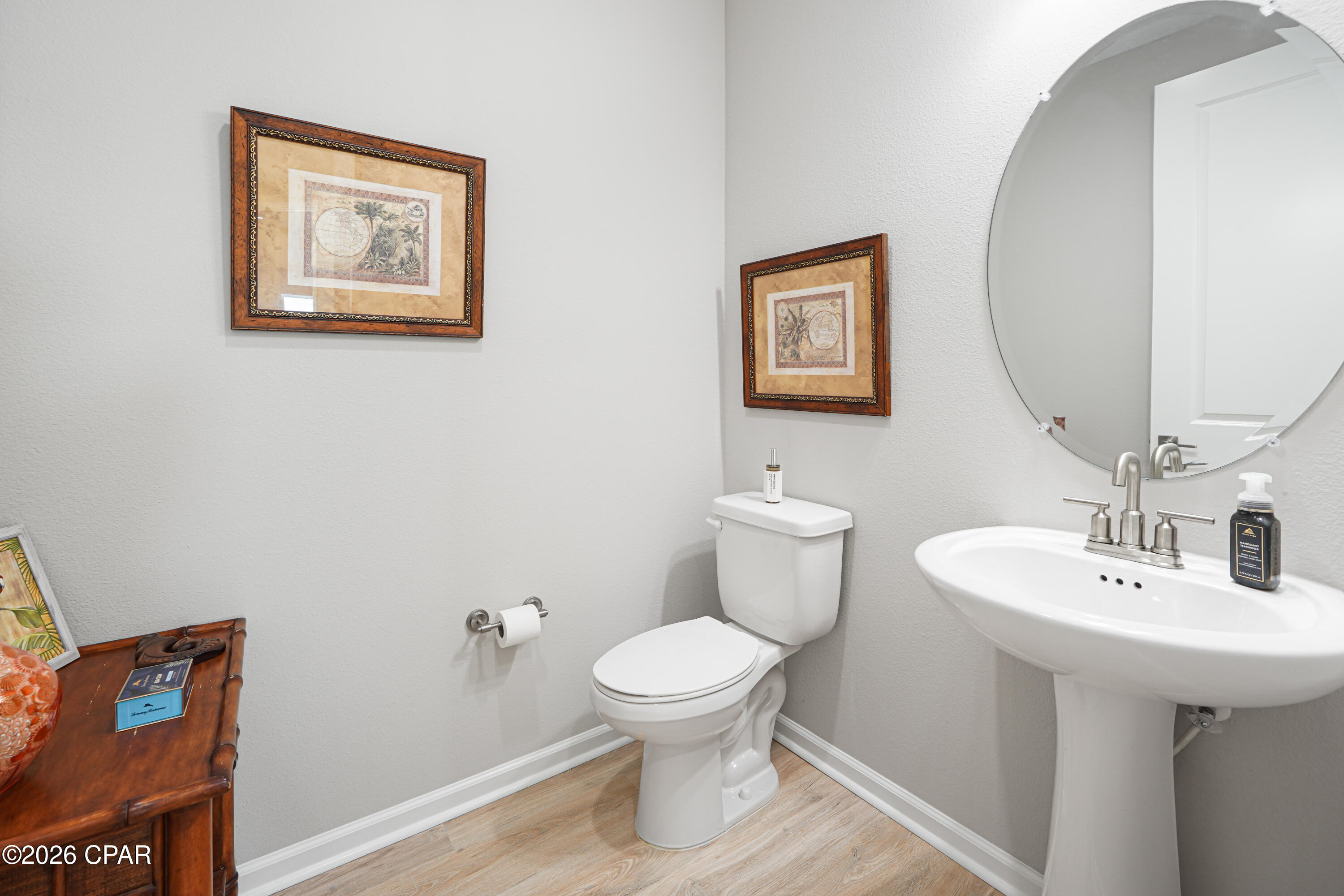 Bathroom with a white pedestal sink, toilet, large round mirror, and framed artwork.