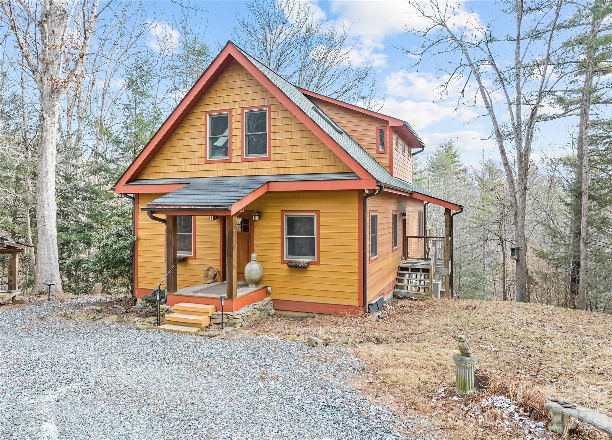 Yellow-orange gabled house with red trim, a covered porch, and a gravel driveway among leafless trees.