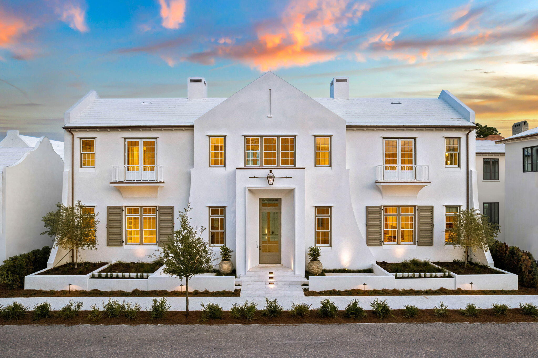 White two-story home with paired balconies and warmly lit windows beneath a colorful sunset sky.