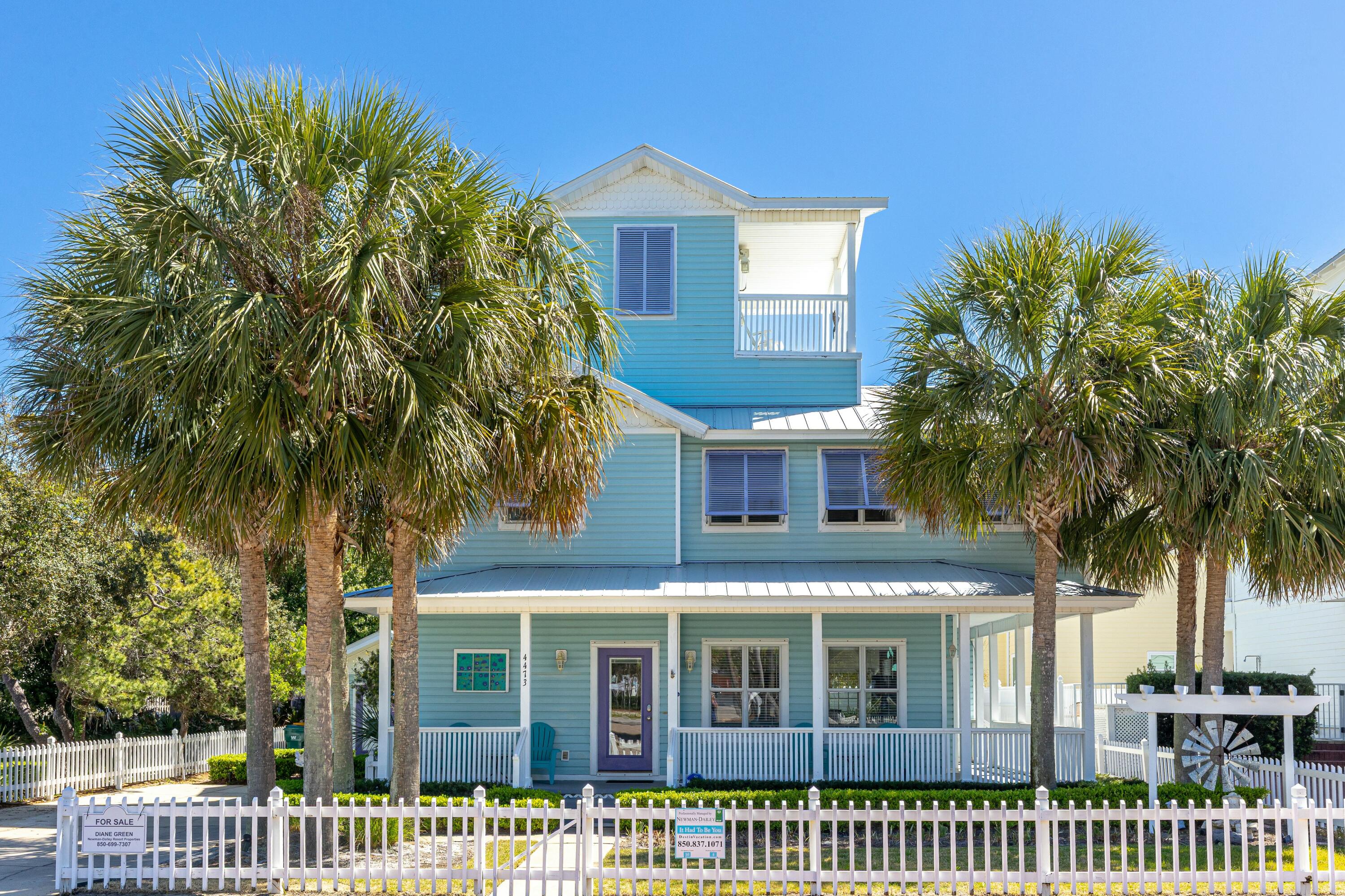 Turquoise coastal-style house with a covered porch, palm trees, and white picket fence.