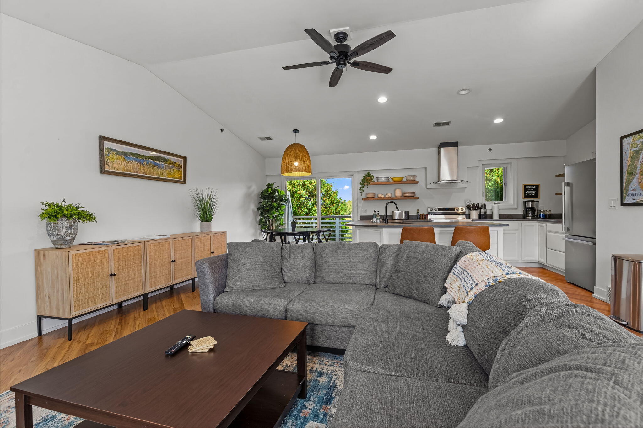 Open-plan living room and kitchen with gray sectional, white cabinetry, and leafy balcony views.