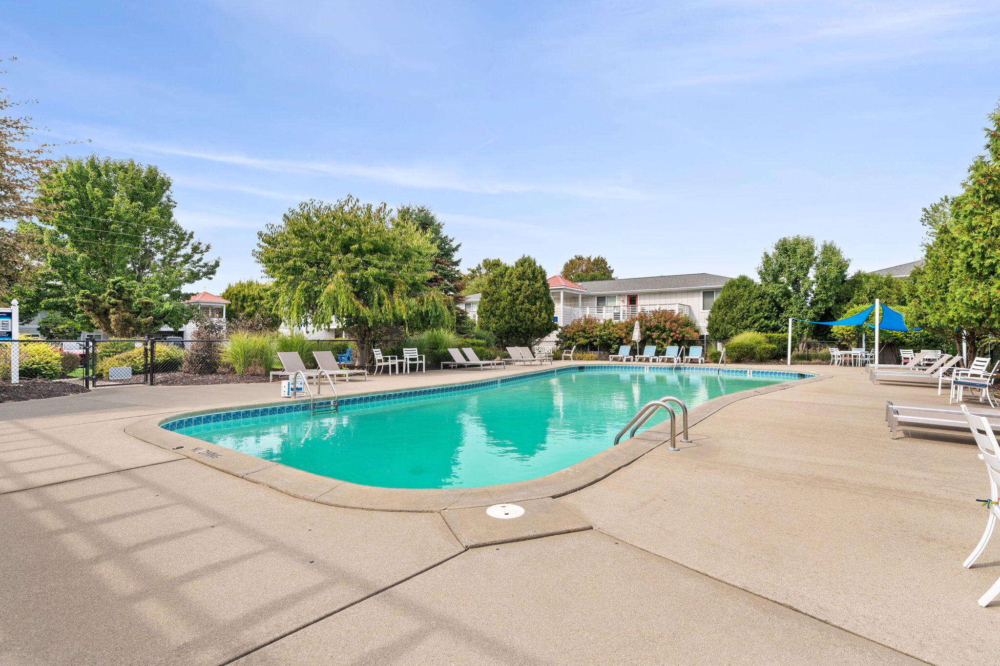 Outdoor swimming pool with lounge chairs and a blue shade sail beside landscaped grounds.