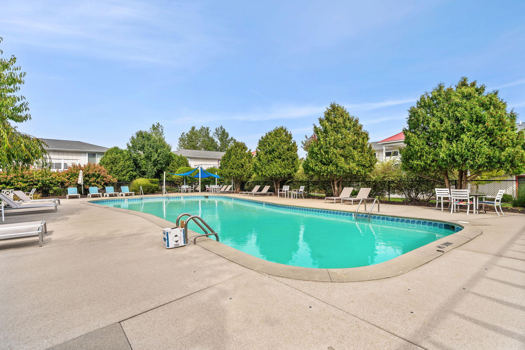 Outdoor swimming pool surrounded by lounge chairs, patio seating, and mature trees.