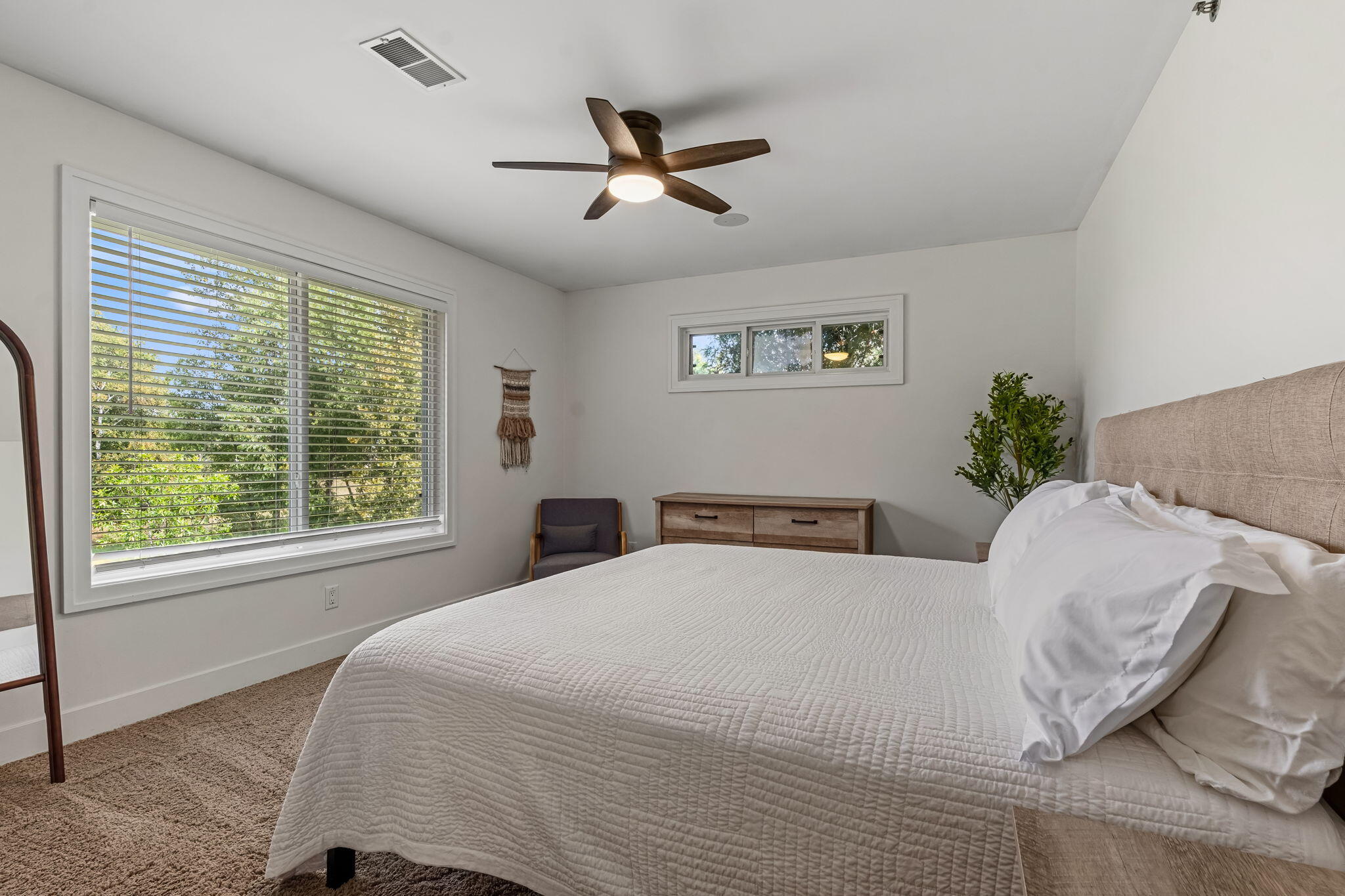 Bedroom with a white quilted bed, wood dresser, and leafy views through large windows.
