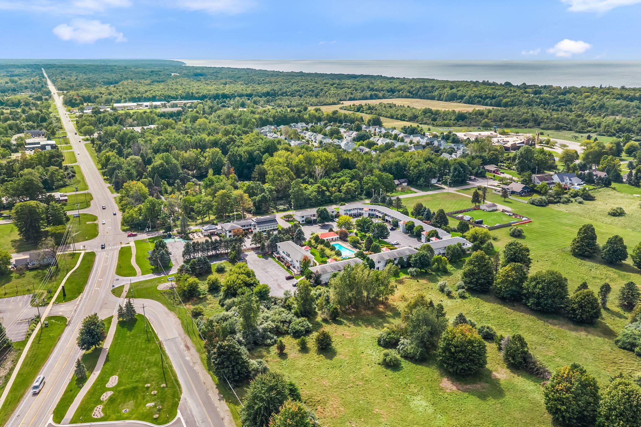Aerial view of a roadside property complex with a turquoise pool, open grounds, and distant water.