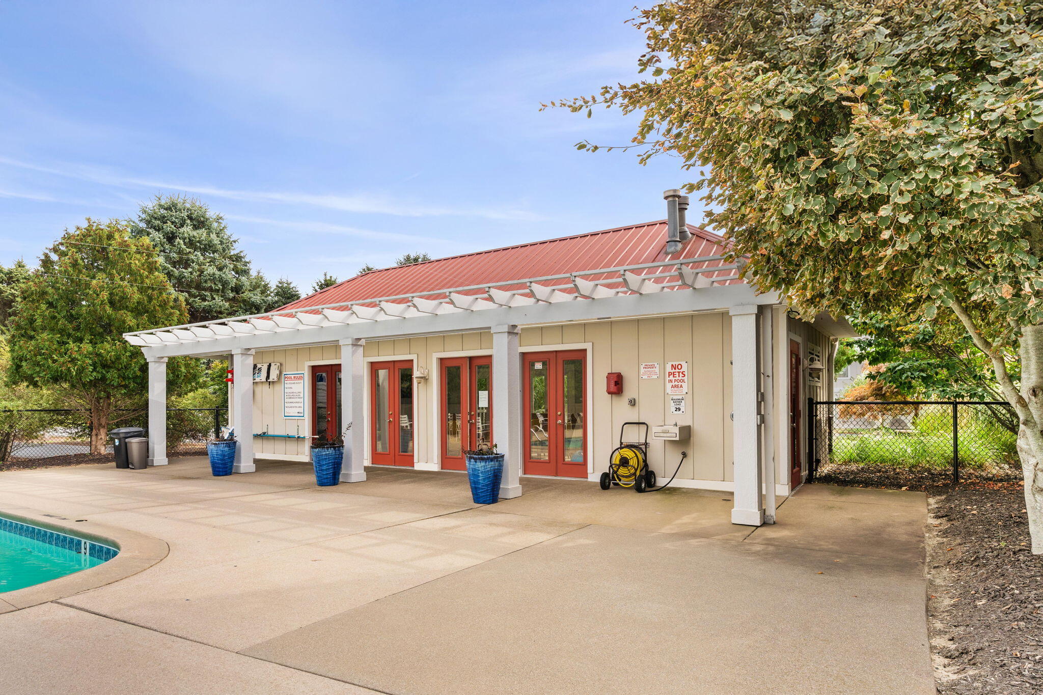 Poolside building with a red metal roof, white pergola, red-framed doors, and concrete deck beside the swimming pool.