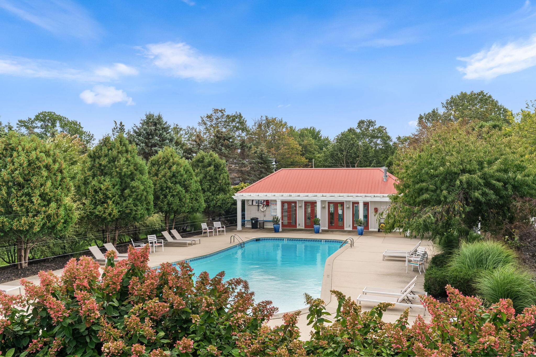 Outdoor pool beside a red-roofed pool house, with patio seating and flowering shrubs.