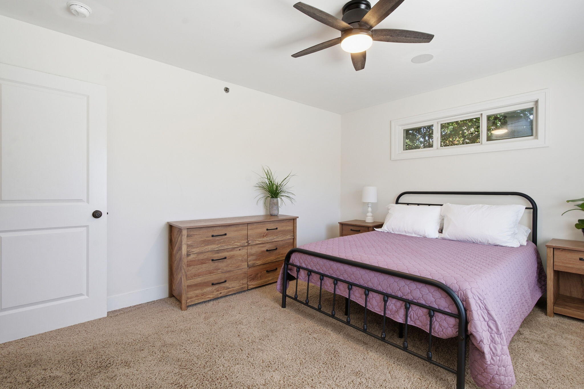 Bedroom with a black metal bed, mauve quilt, wooden dresser, and high horizontal window.