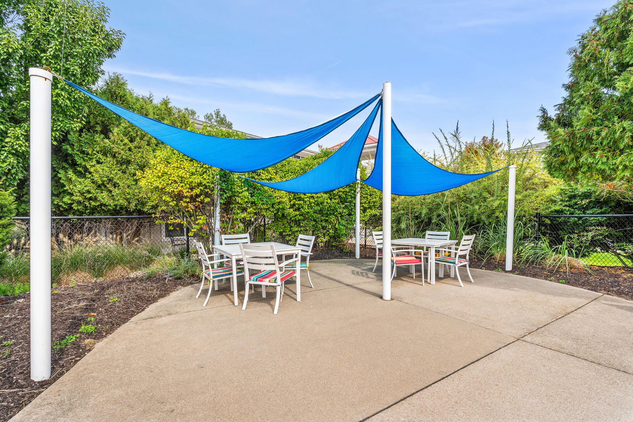 Patio dining area with blue shade sails, white tables and chairs, and landscaped borders.