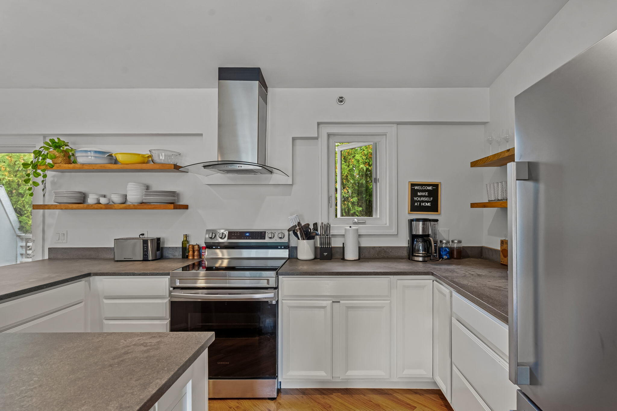 Bright white kitchen with stainless steel appliances, open wood shelves, and a window overlooking greenery.