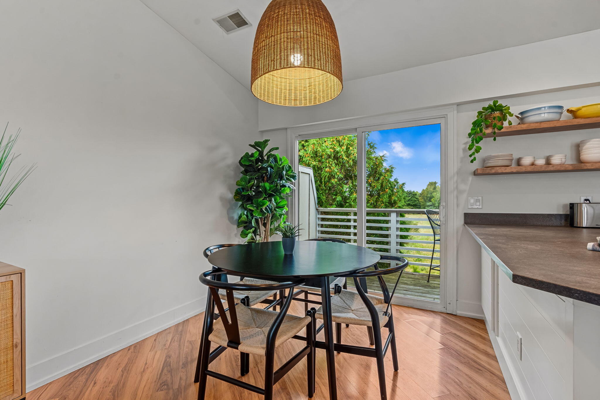 Dining nook with a round black table and woven chairs beside sliding doors to a tree-lined deck.