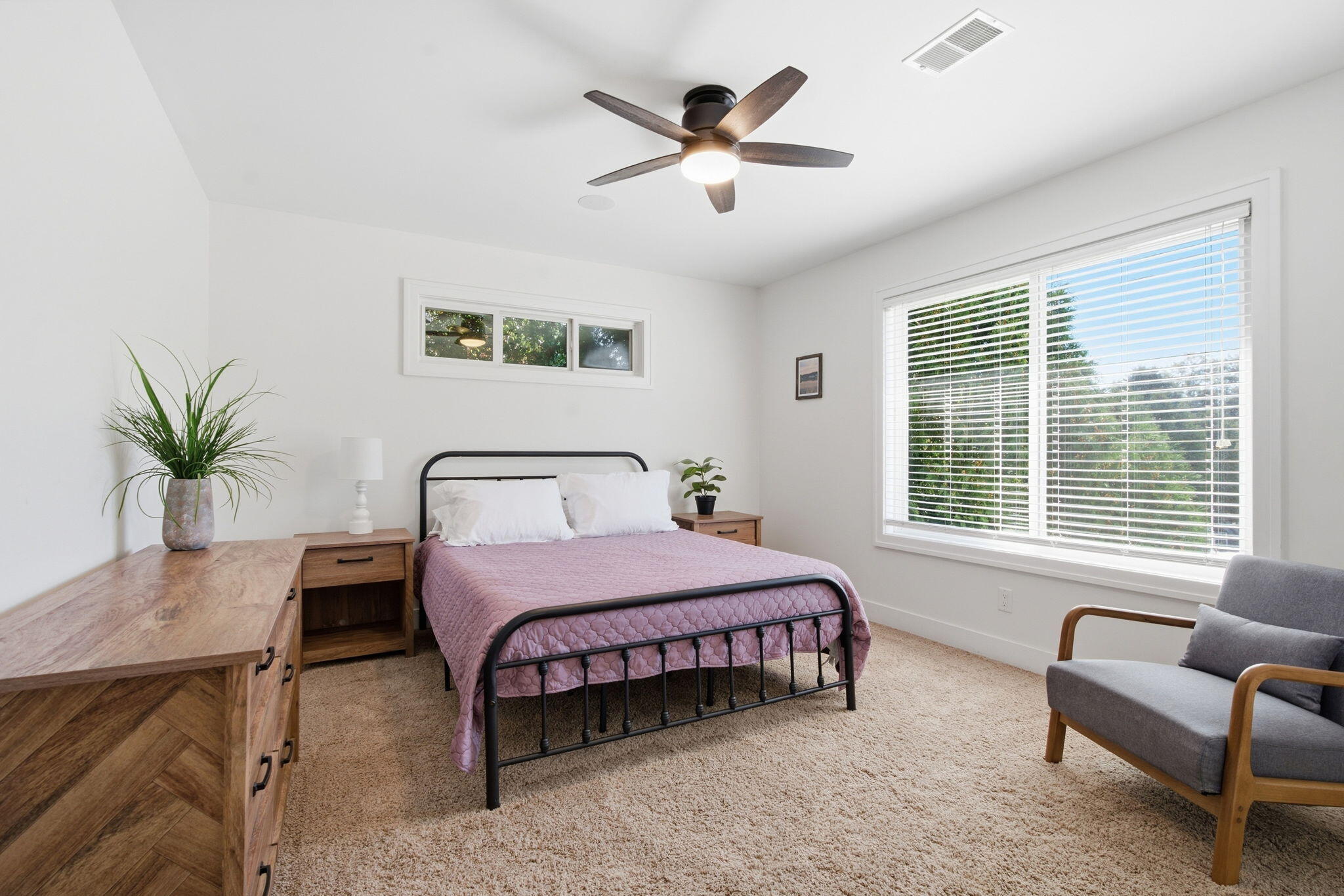 Bedroom with a black metal bed, mauve quilt, wood furniture, ceiling fan, and large window.