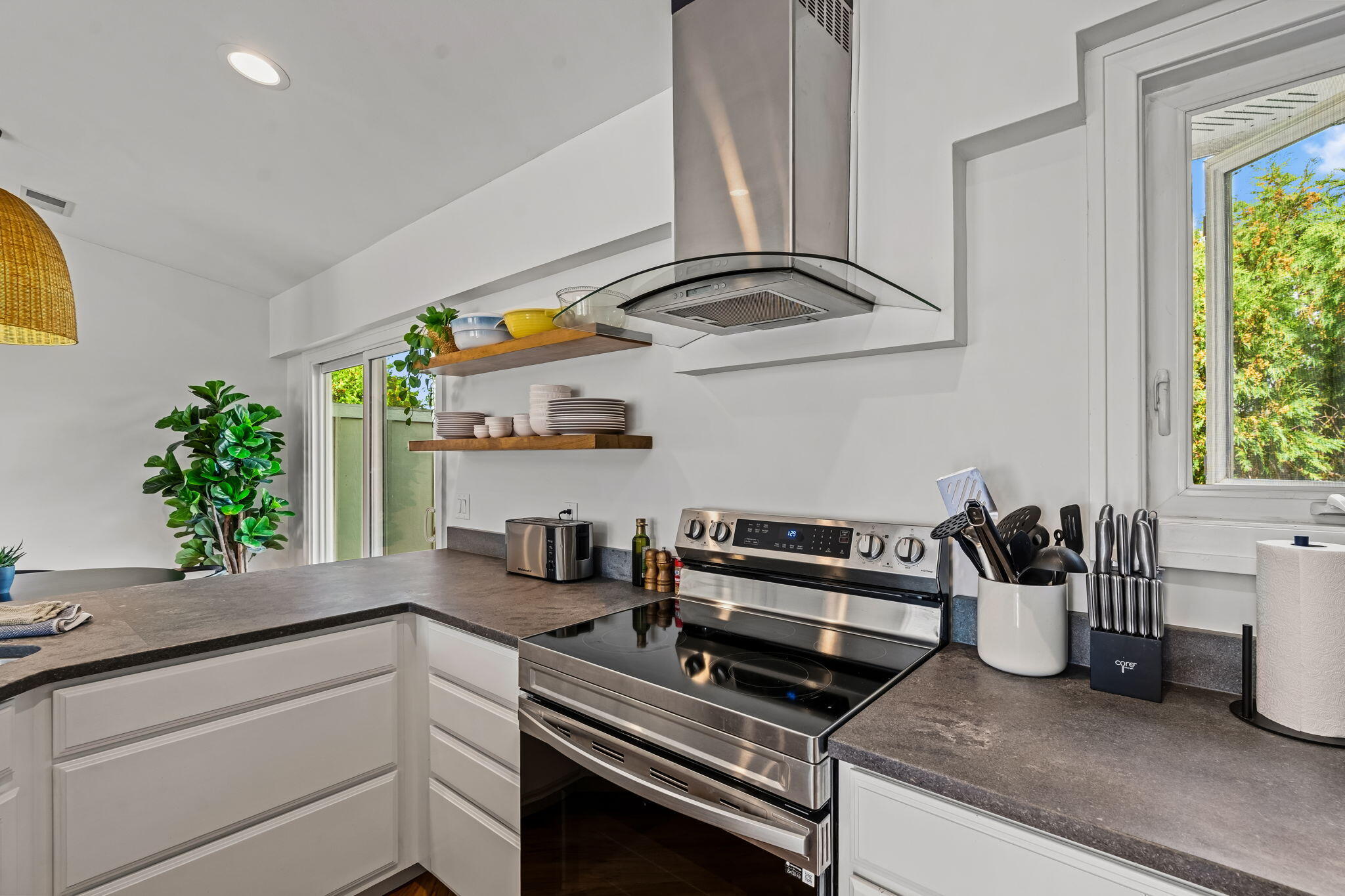 Bright kitchen with stainless steel range, glass hood, dark counters, and floating wood shelves.