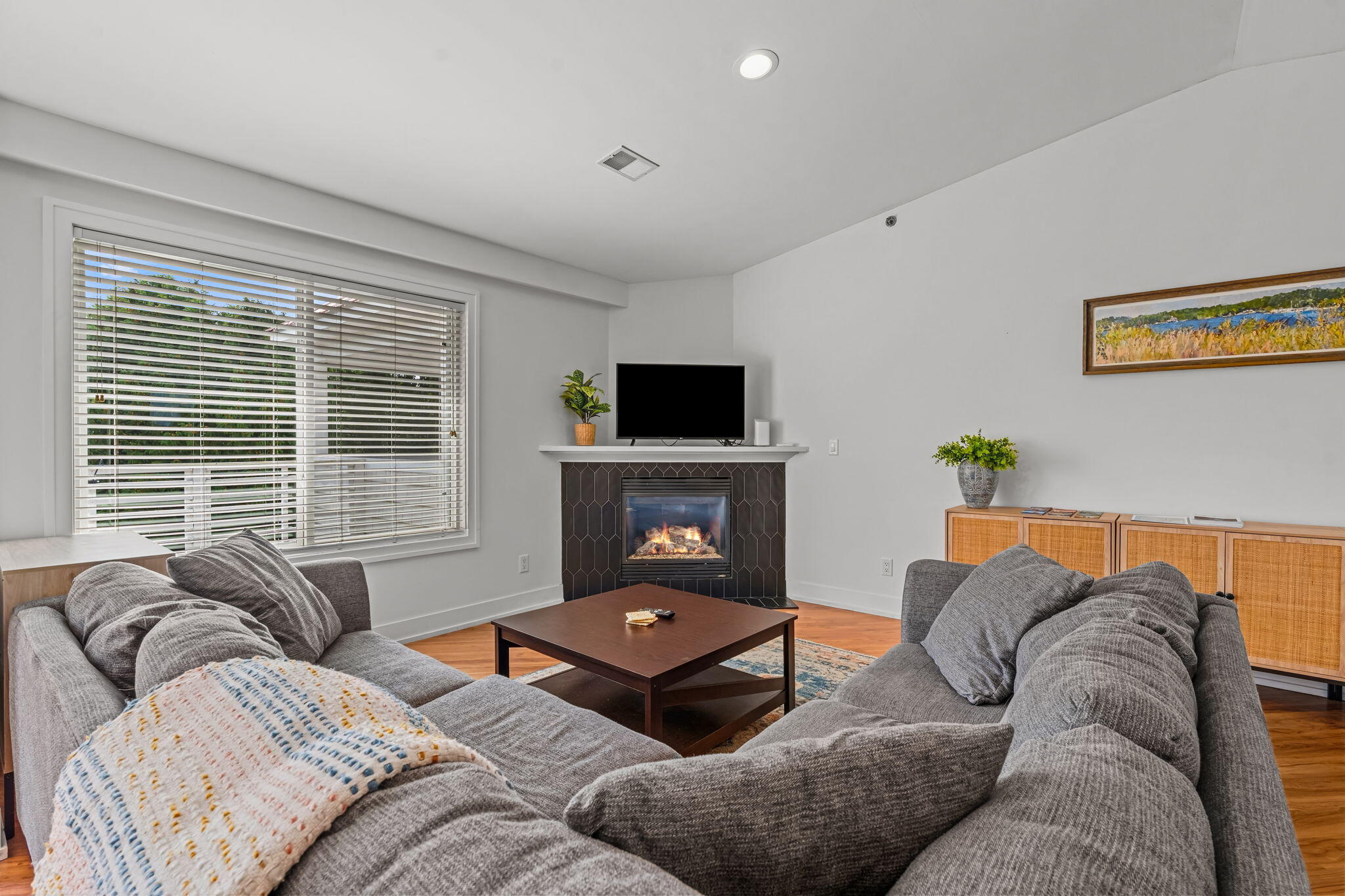 Living room with gray sofas, a lit tiled fireplace, hardwood floors, and a large window with blinds.