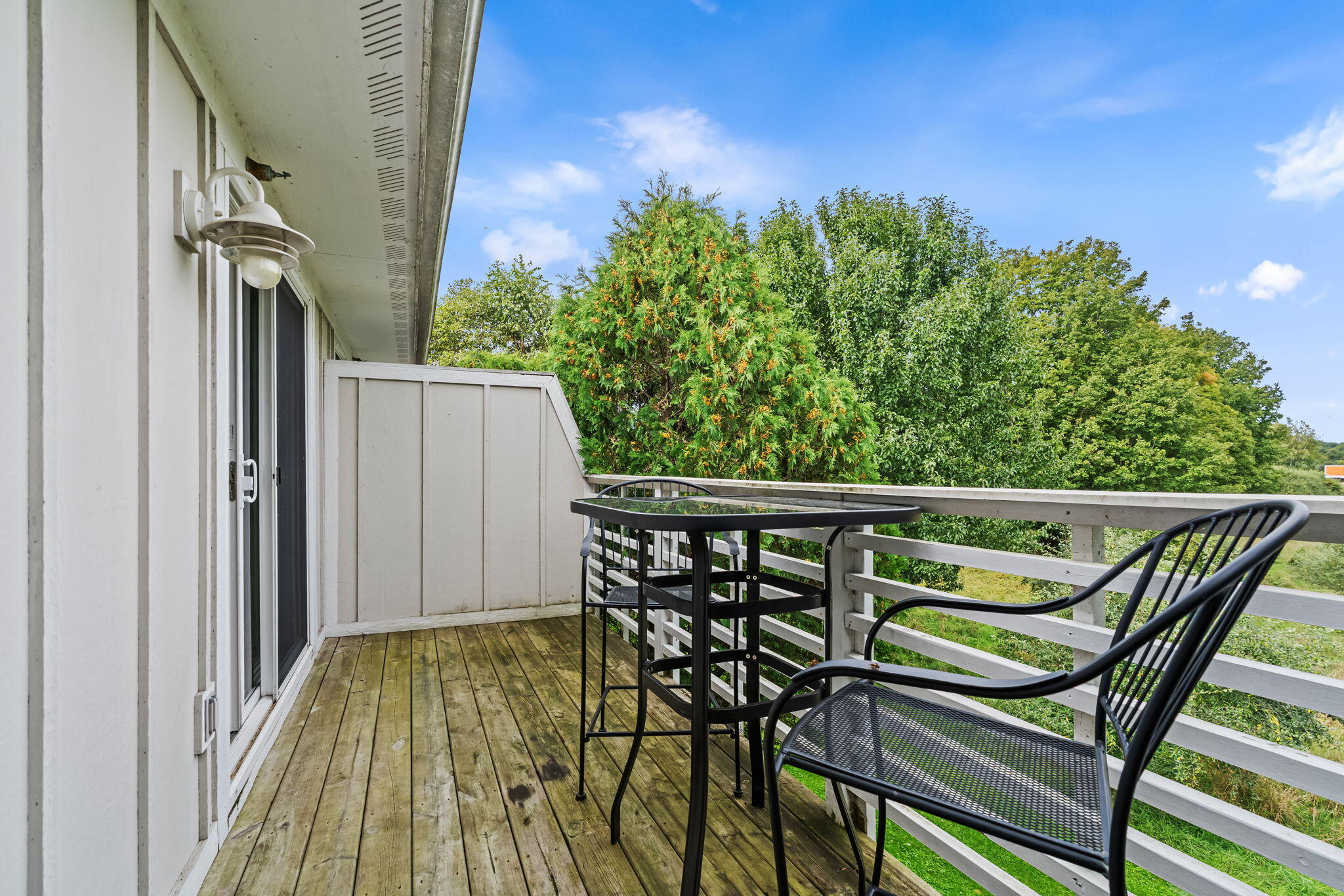 Wooden balcony with black metal patio furniture overlooking leafy trees.