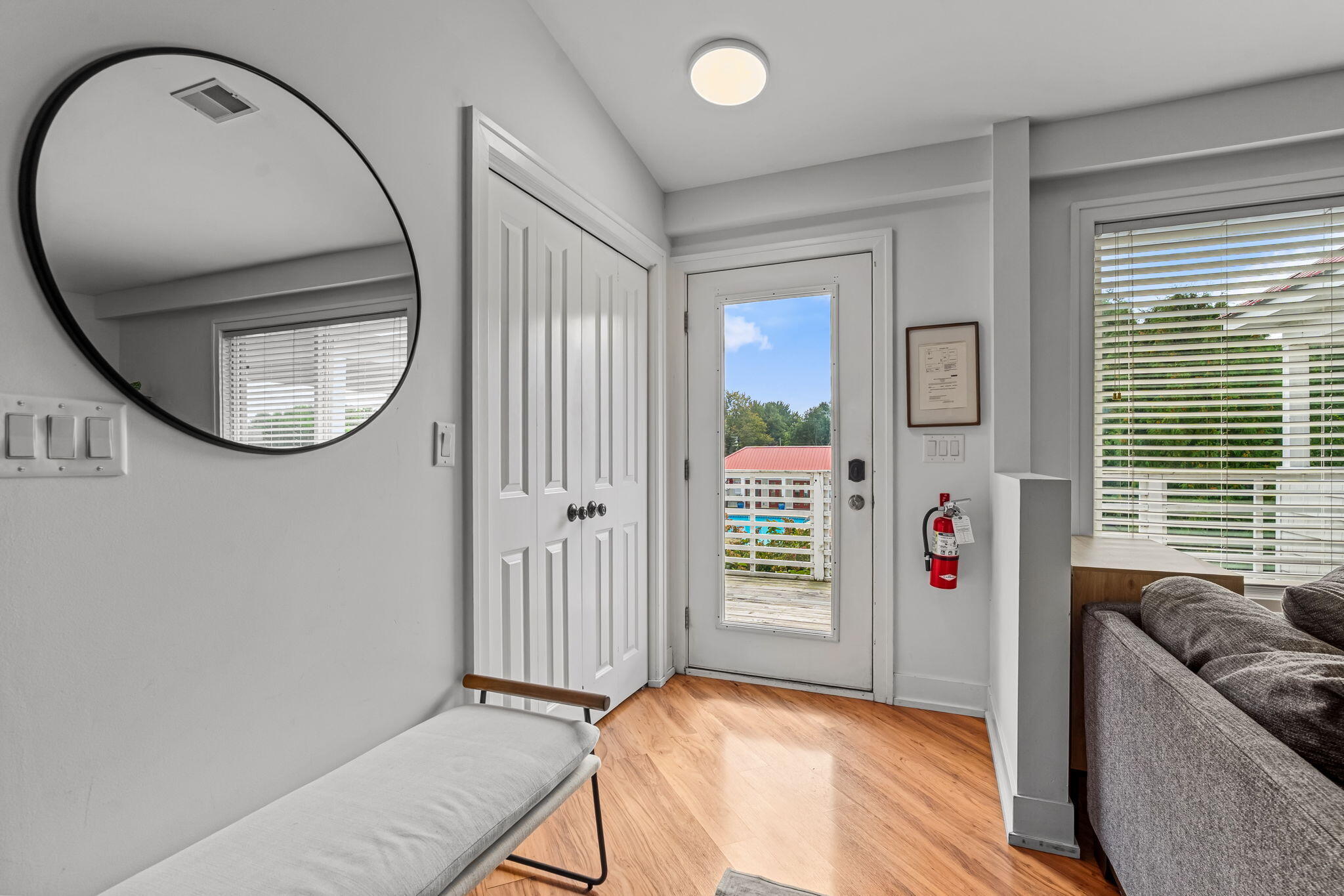 Bright entryway with a glass-panel front door, double closets, round mirror, bench, and gray sofa.