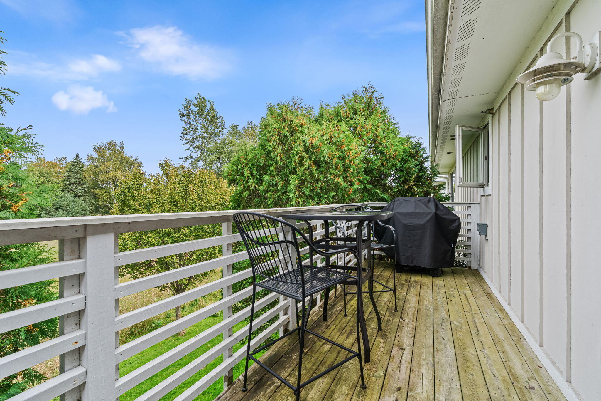 Wooden deck with black dining furniture and a covered grill, bordered by white railings and leafy trees.