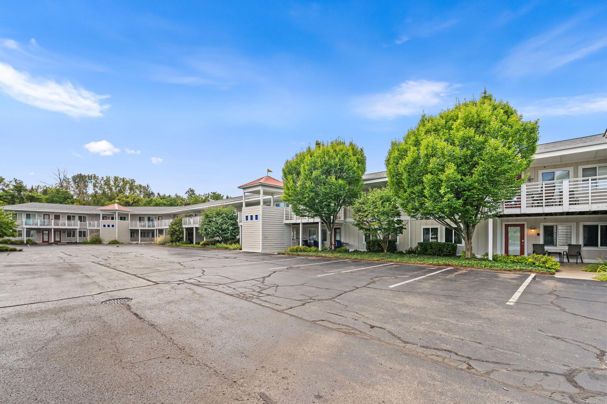 Two-story multifamily building with white balconies, leafy trees, and a large paved parking lot.