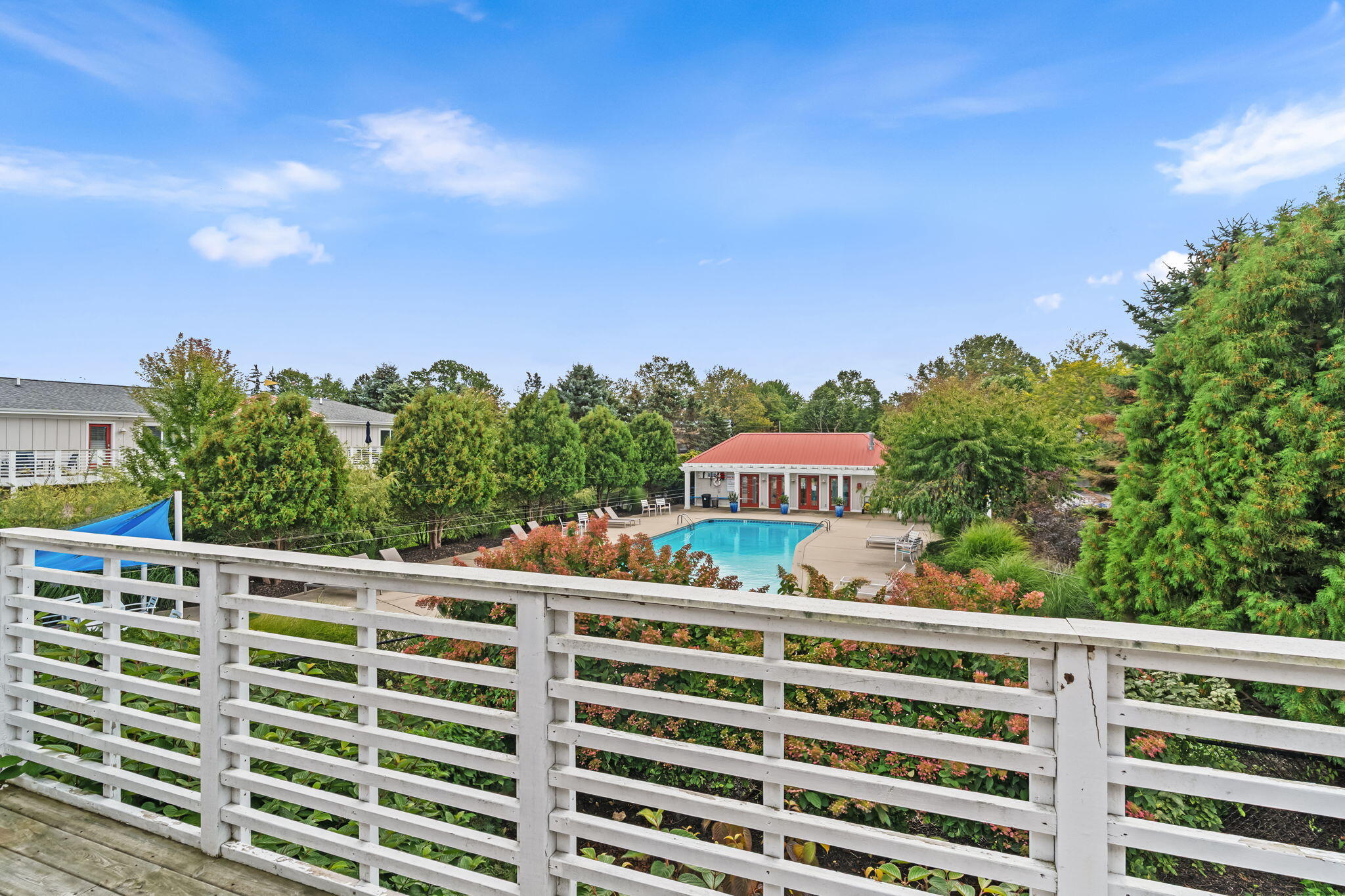 Outdoor pool and red-roofed pool building amid landscaped grounds, viewed over a white deck railing.