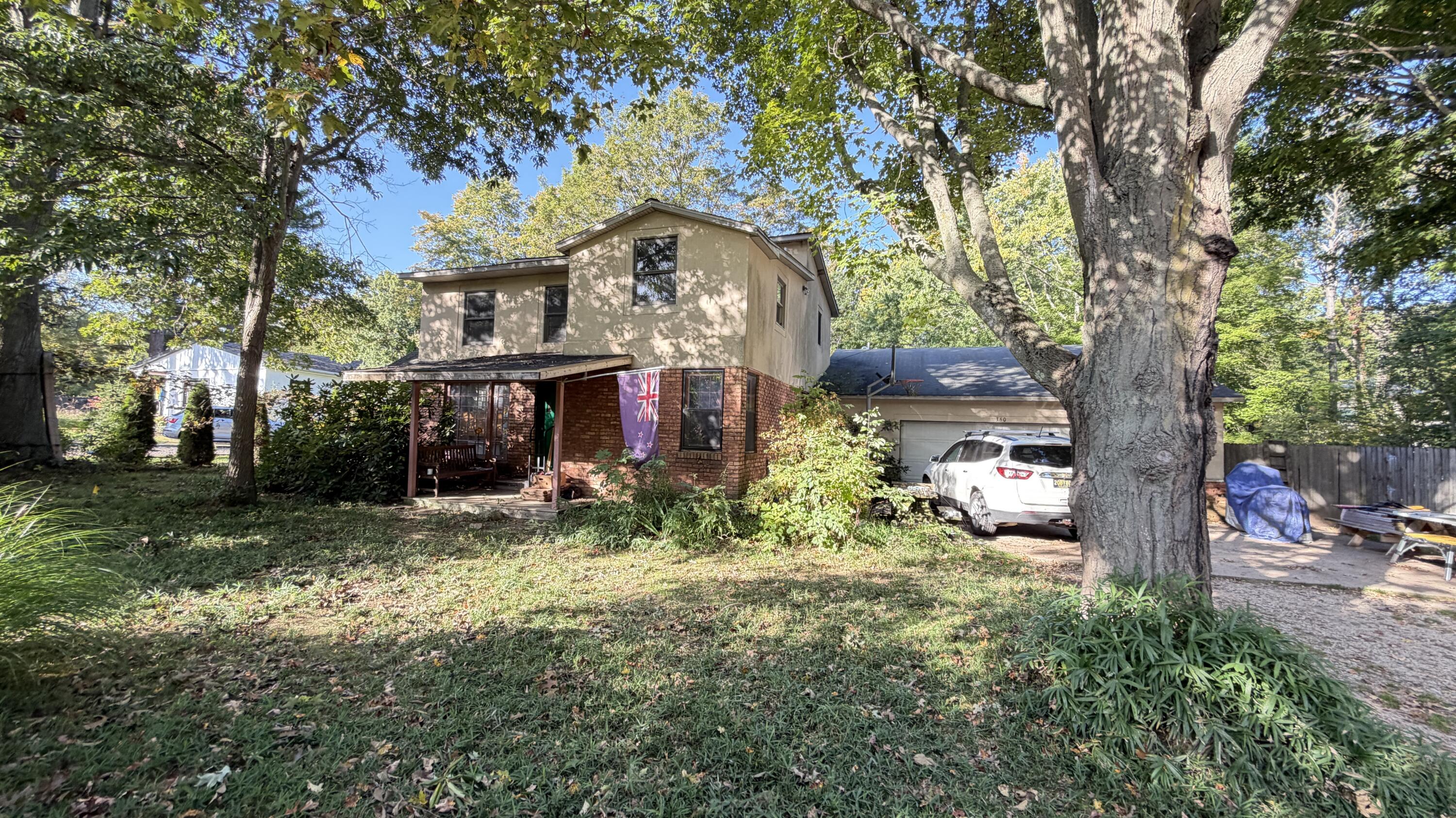 Two-story beige and brick house with a covered porch, attached garage, and large tree in the front yard.